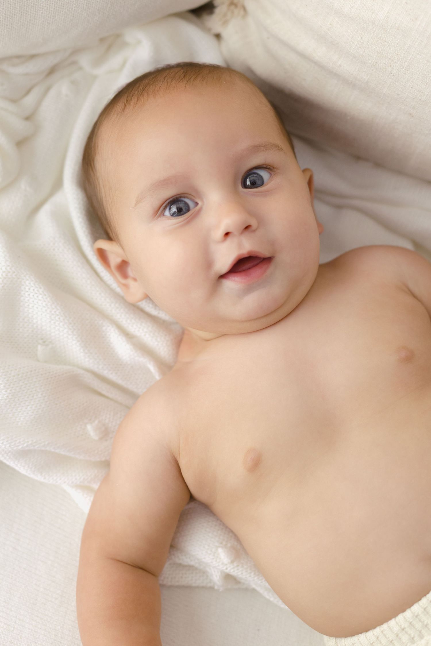 Adorable baby with blue eyes lying on a soft white blanket, looking up with a curious and happy expression.