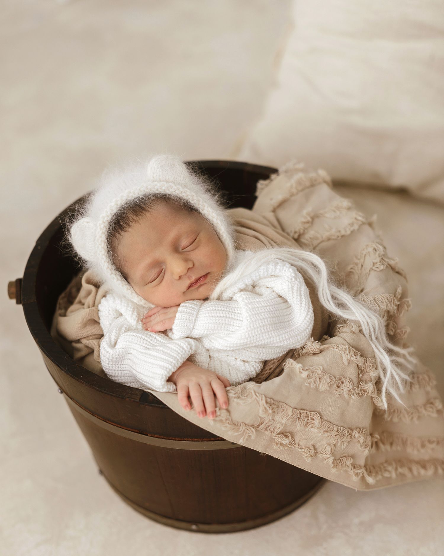 Newborn baby in white knit outfit and bear ear bonnet sleeping in rustic wooden bucket.
