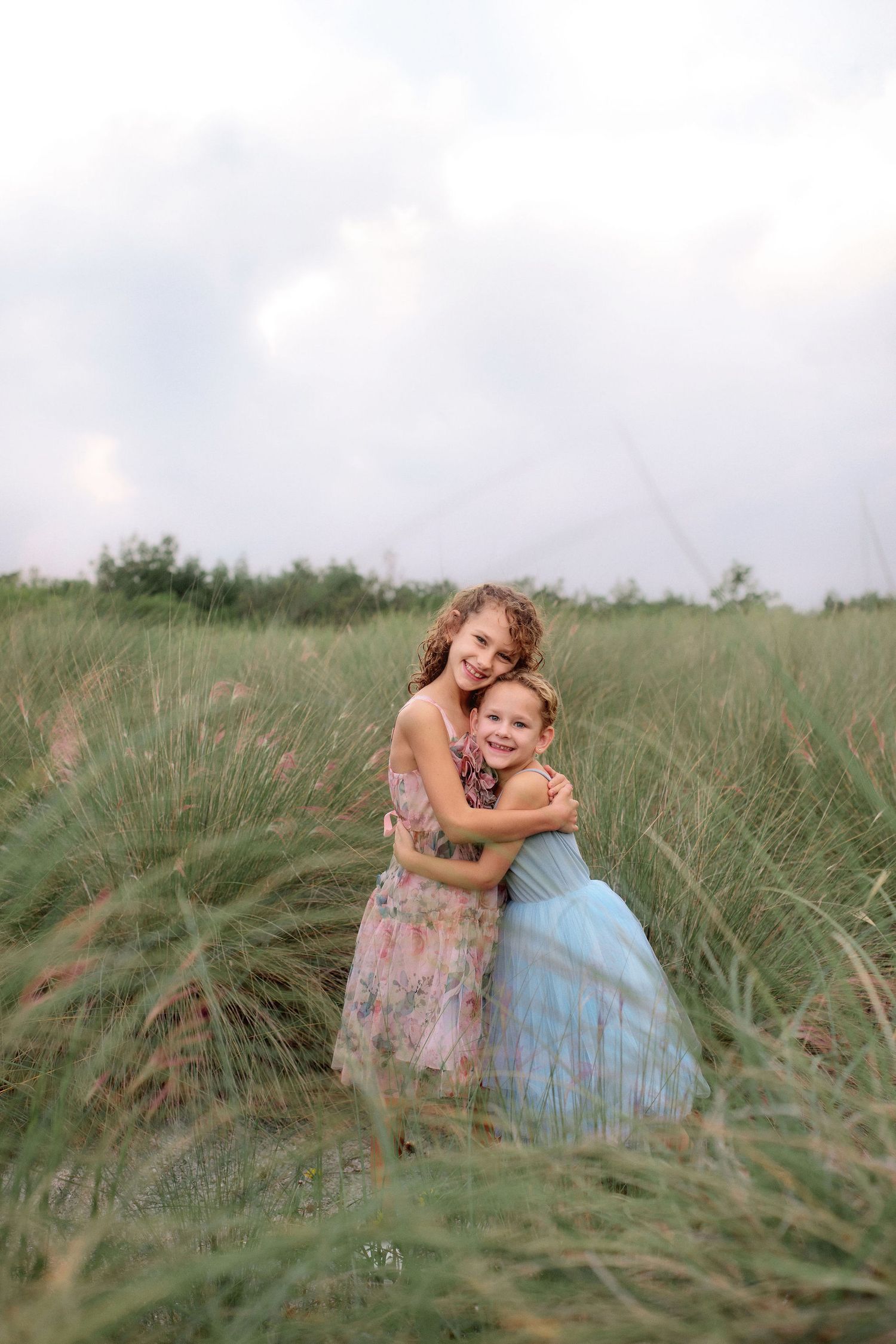 Two young girls in pastel floral and blue tulle dresses hug and smile in a tall green grass field under a soft cloudy sky.