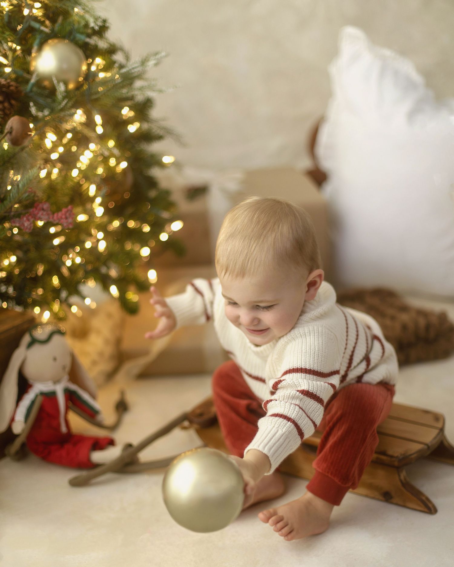 A baby in a white and red sweater sits on a wooden sled reaching for a gold ornament near a lit Christmas tree.