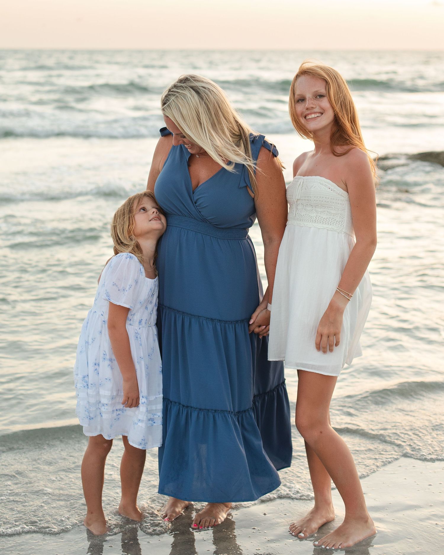 A mother and two daughters pose together on a sandy beach at sunset, wearing blue and white dresses.