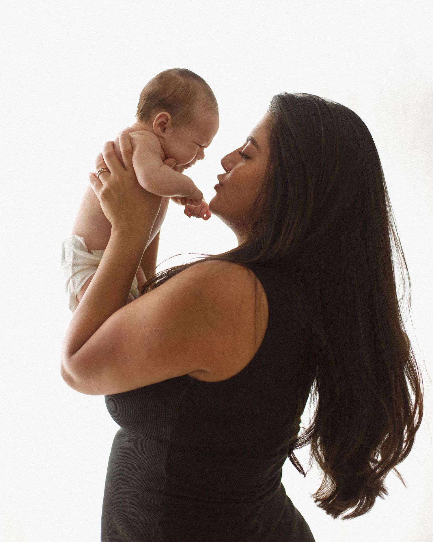 A dark-haired woman in a black top lovingly lifts her smiling newborn baby up close to her face.