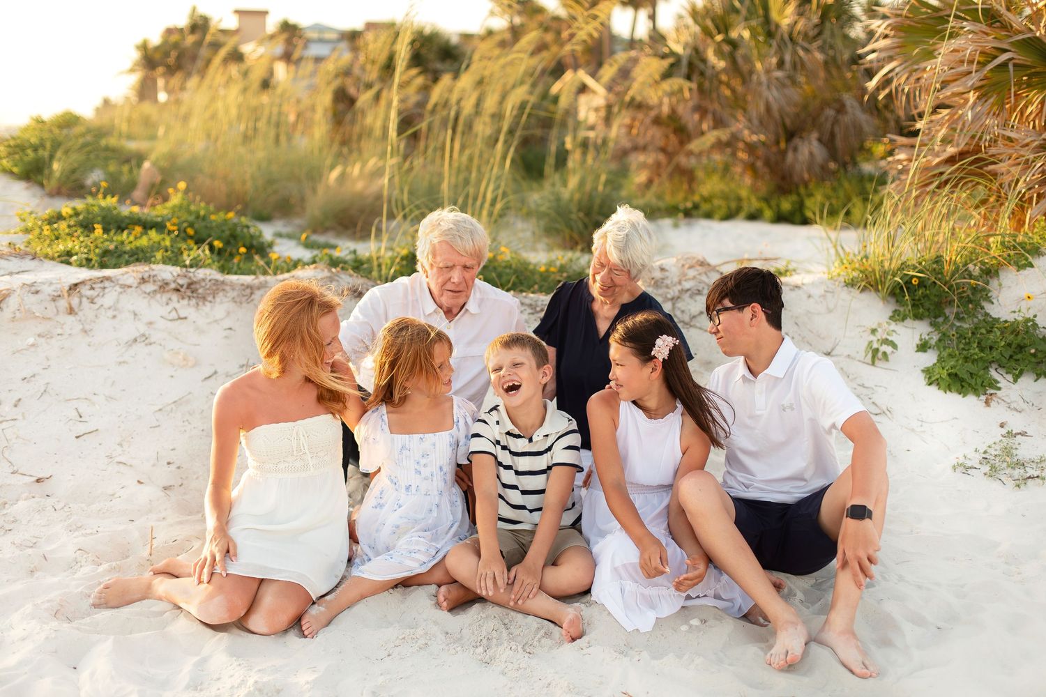 Multi-generational family laughing together on a sandy beach, dressed in white and navy outfits at golden hour.