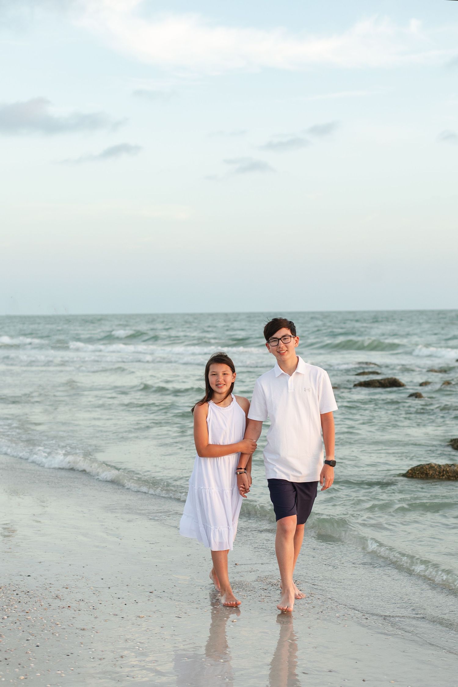 Two young siblings holding hands and walking barefoot along a sandy beach with ocean waves in the background.