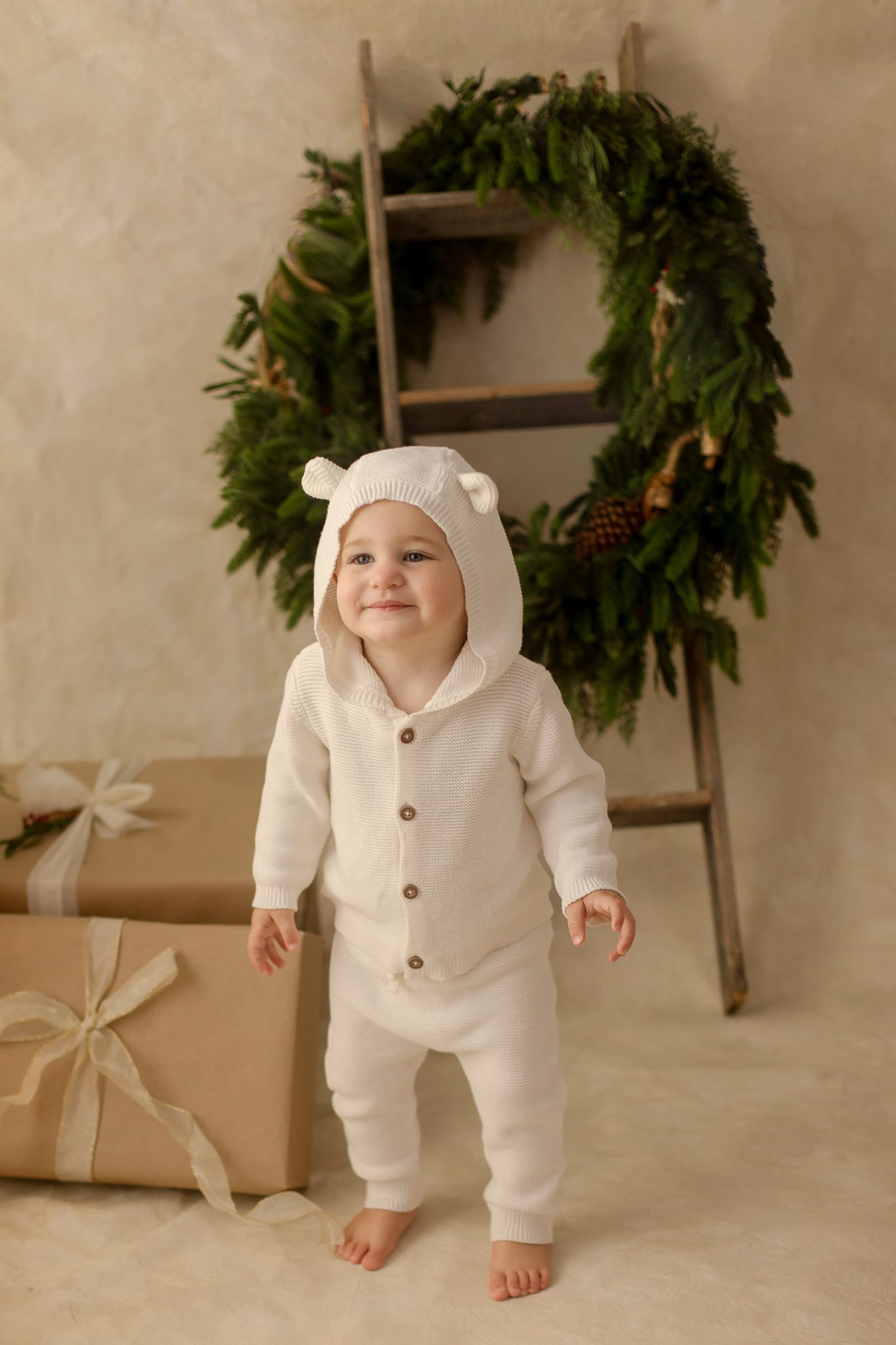 Smiling baby in white bear hoodie romper stands barefoot near Christmas wreath and gifts.