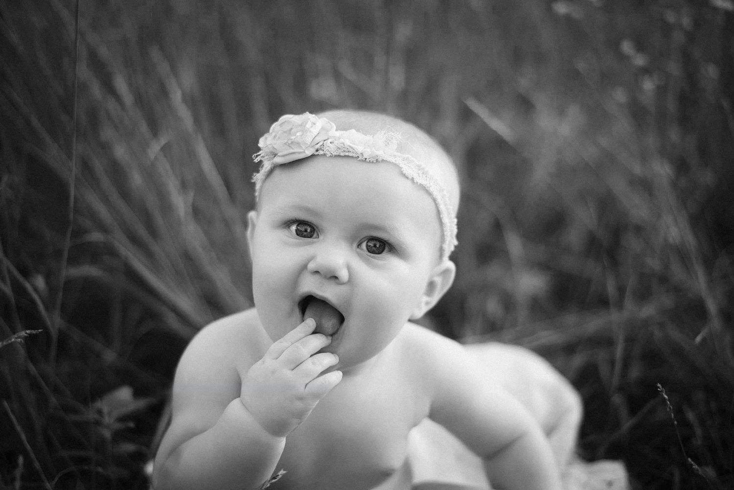 Black and white photo of an adorable baby girl with headband sitting outdoors, chewing on a small object.
