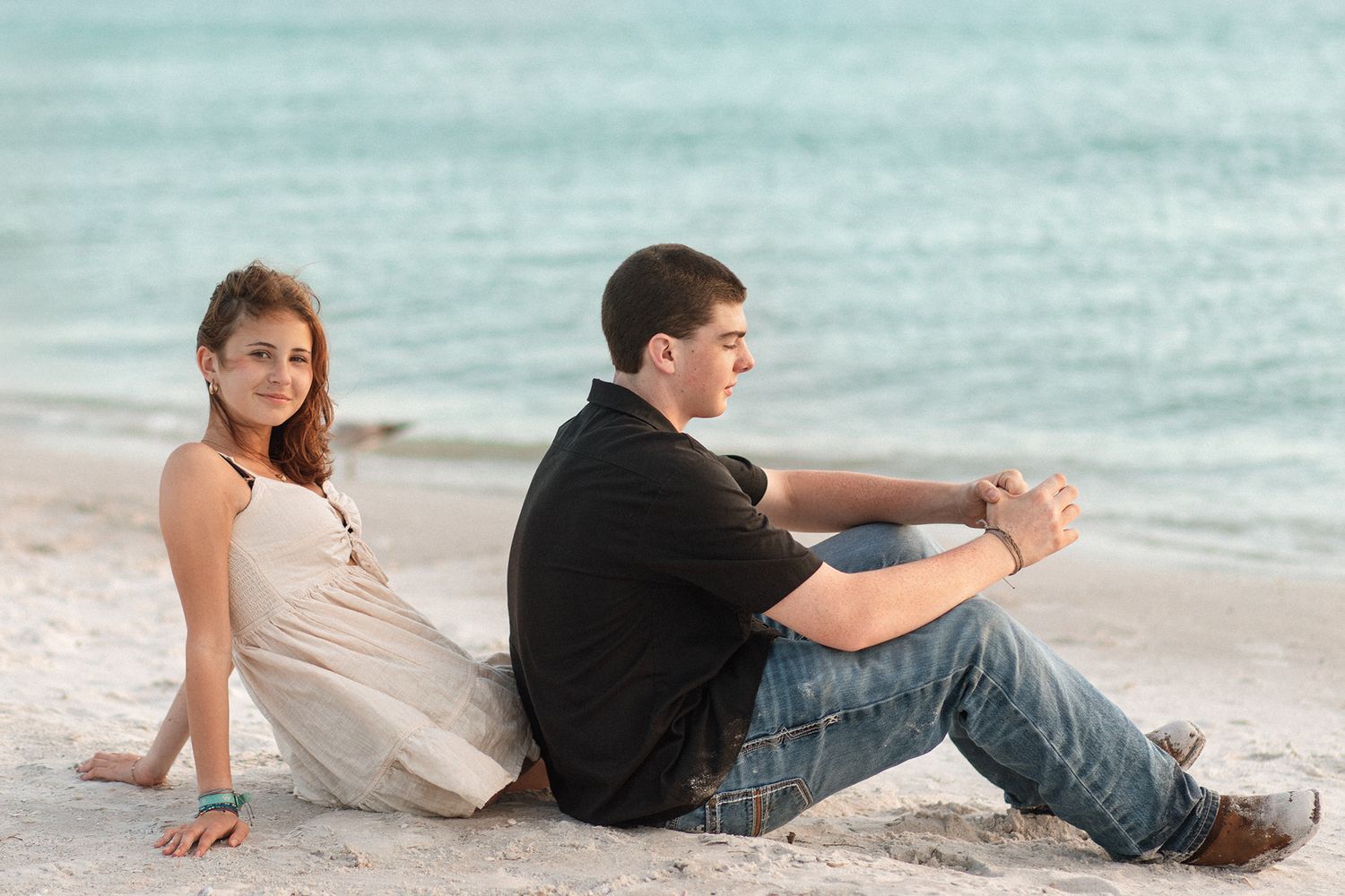 Young couple sitting back to back on a sandy beach with turquoise ocean waves in the background.