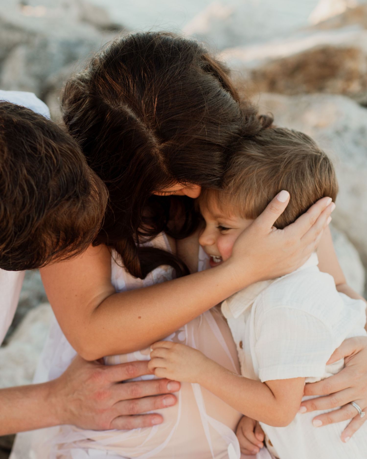 A mother embracing two young children in a warm group hug near rocky terrain, dressed in white outfits.