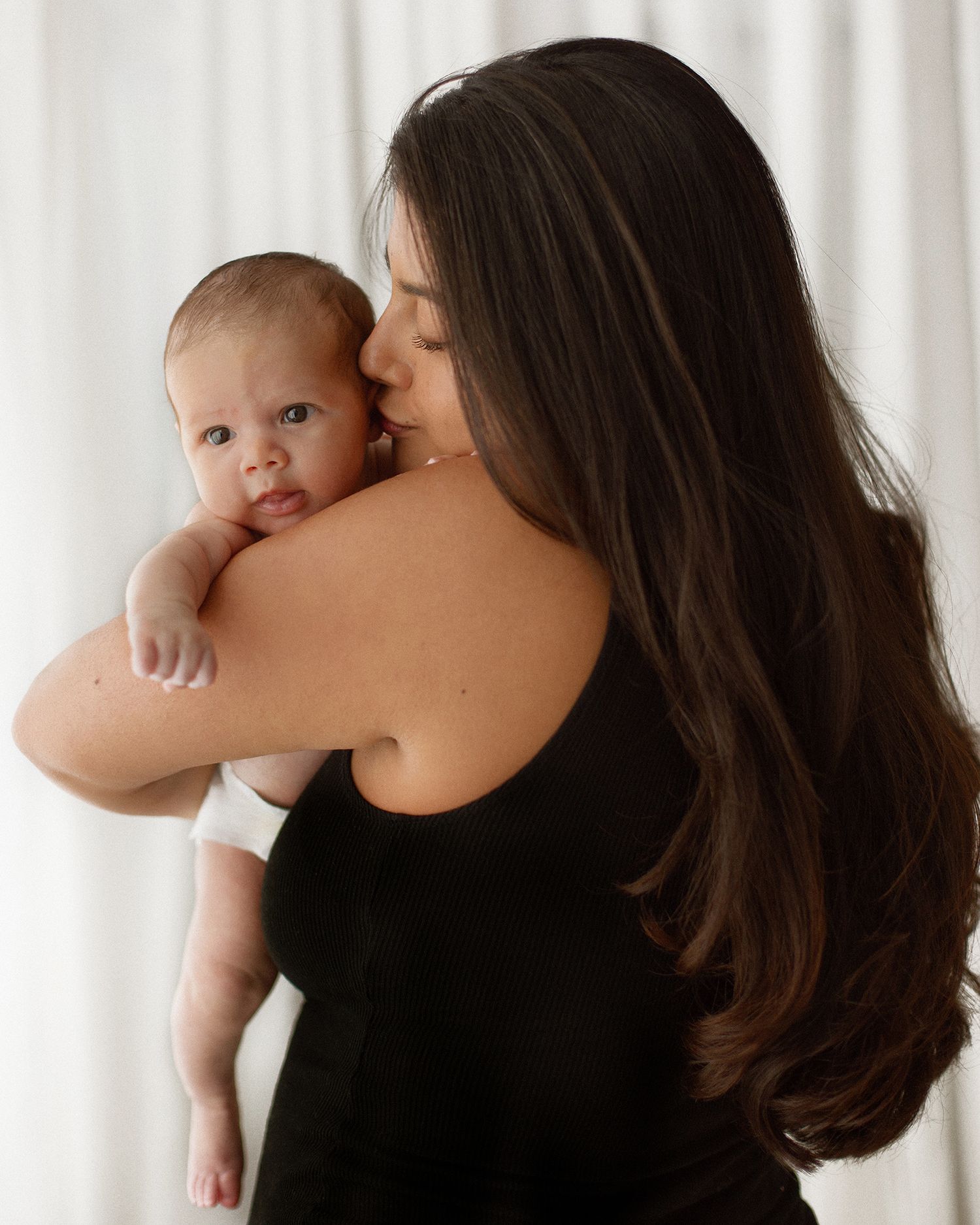 A mother with long dark hair in a black dress kisses her newborn baby while holding them against her shoulder.