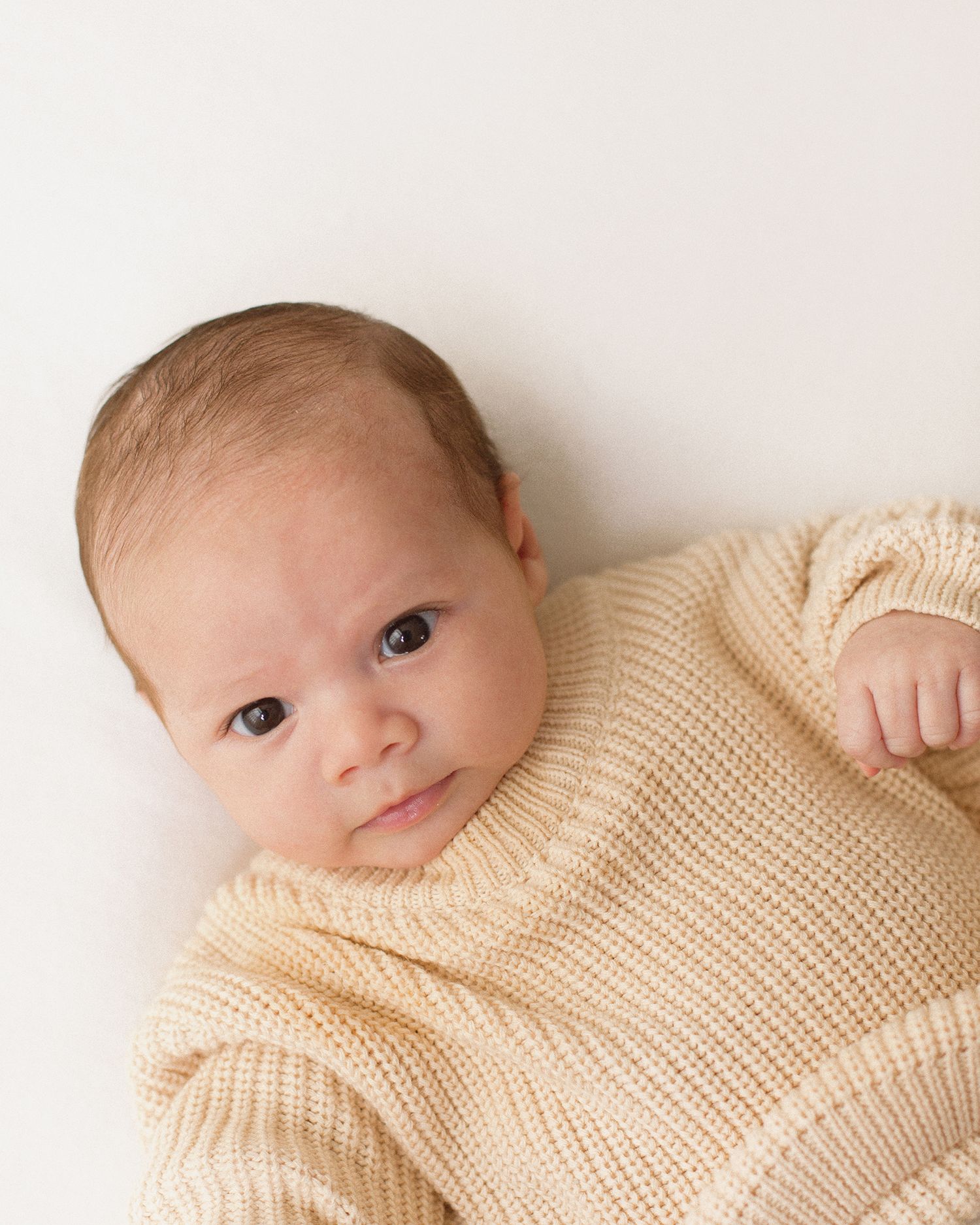 A newborn baby in a cream knit sweater lies on a white background, looking up with curious wide eyes.