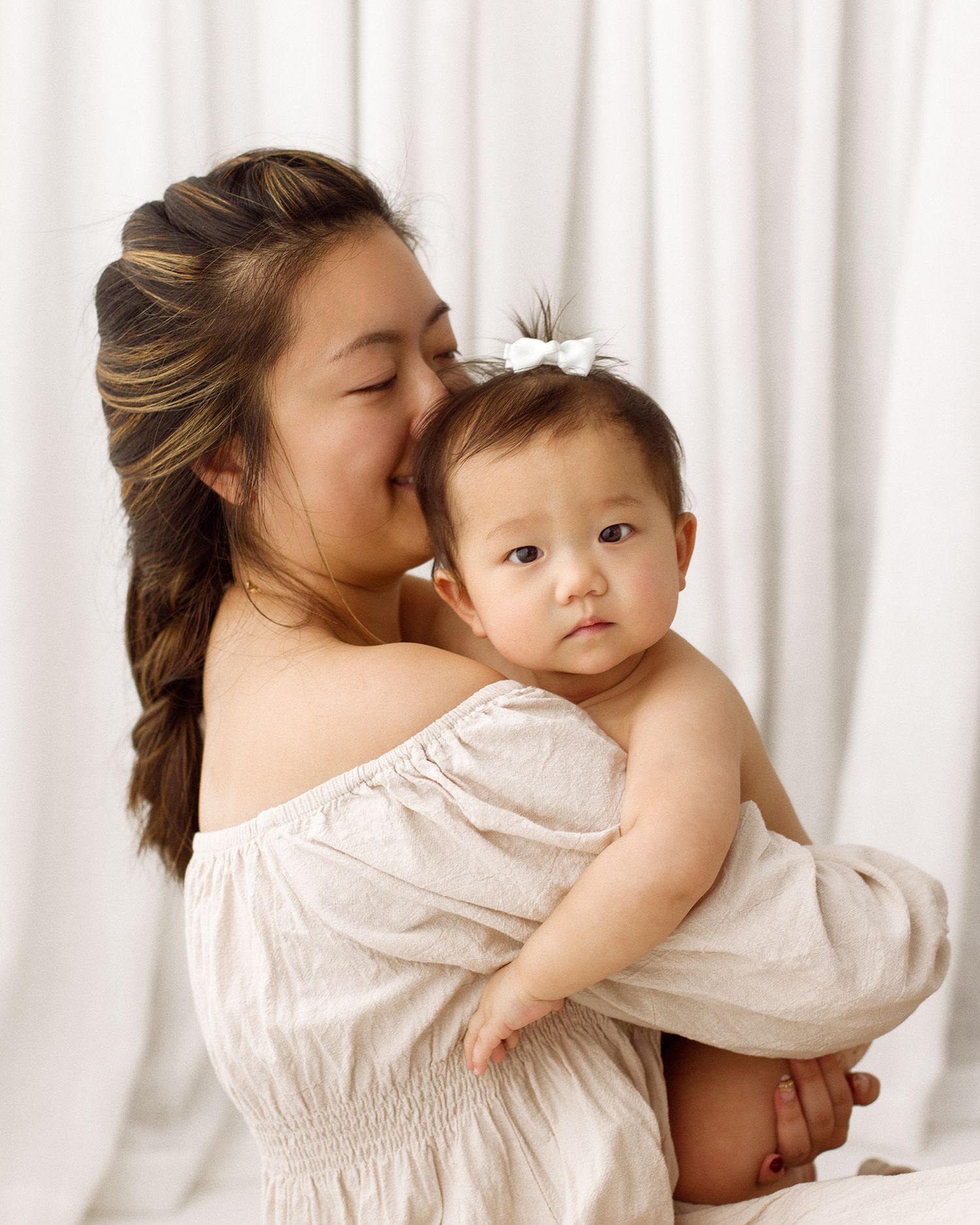 A mother with a braided ponytail kisses her baby's cheek while holding her close, in a soft studio portrait with white drapes.