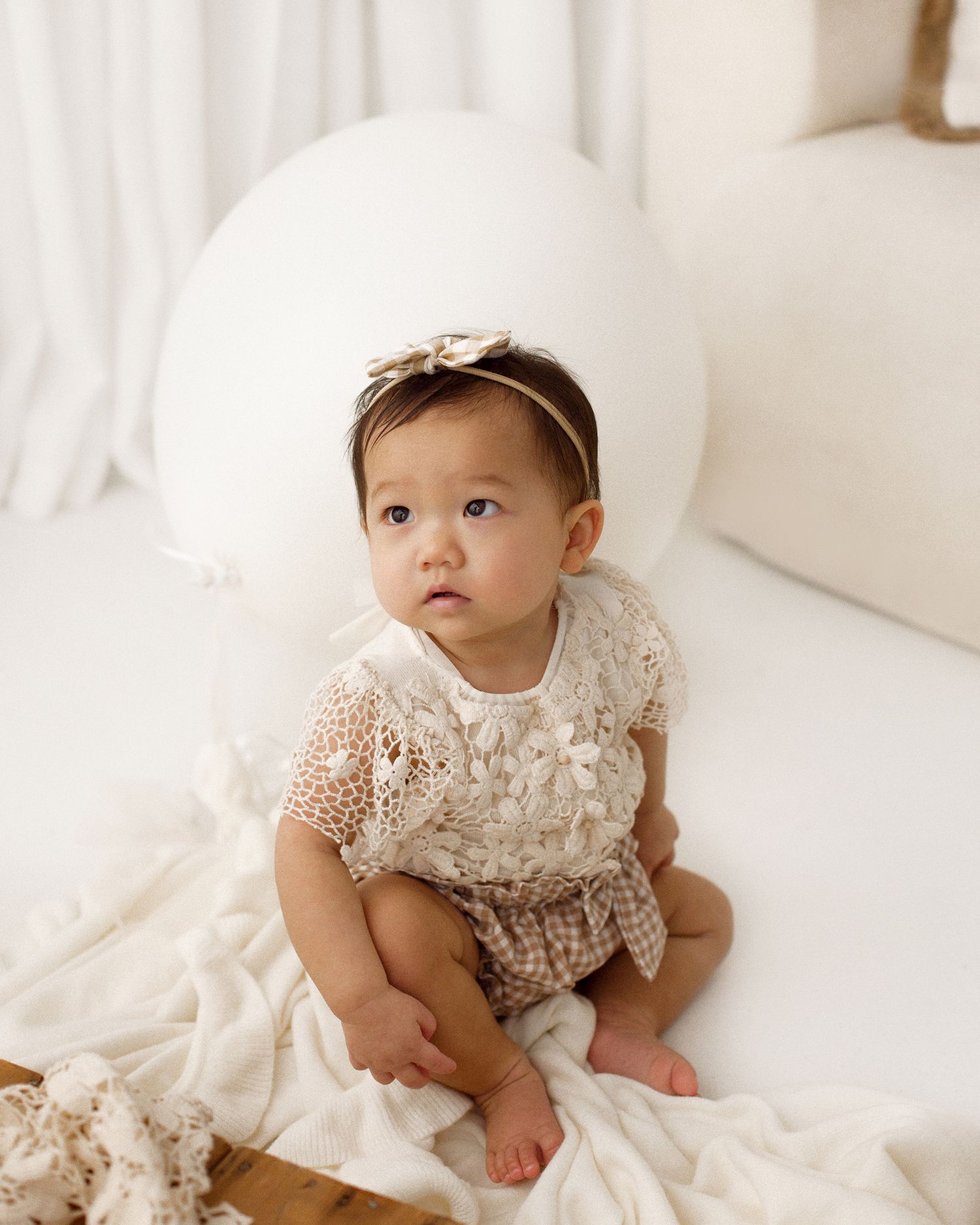 Baby girl in a cream lace romper and bow headband sits on a soft white blanket in a bright, airy studio portrait.