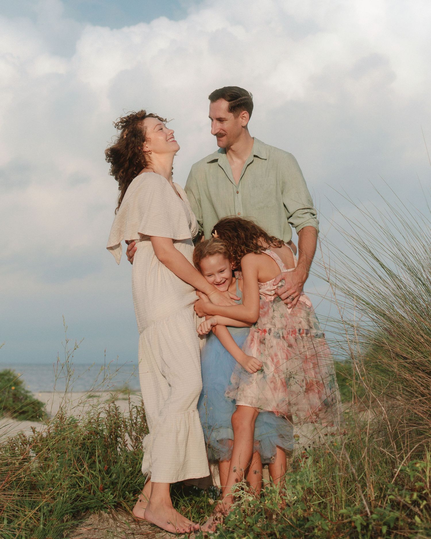 Smiling family of four hugging on a grassy beach dune at sunset, with parents and two young daughters in light summer outfits.