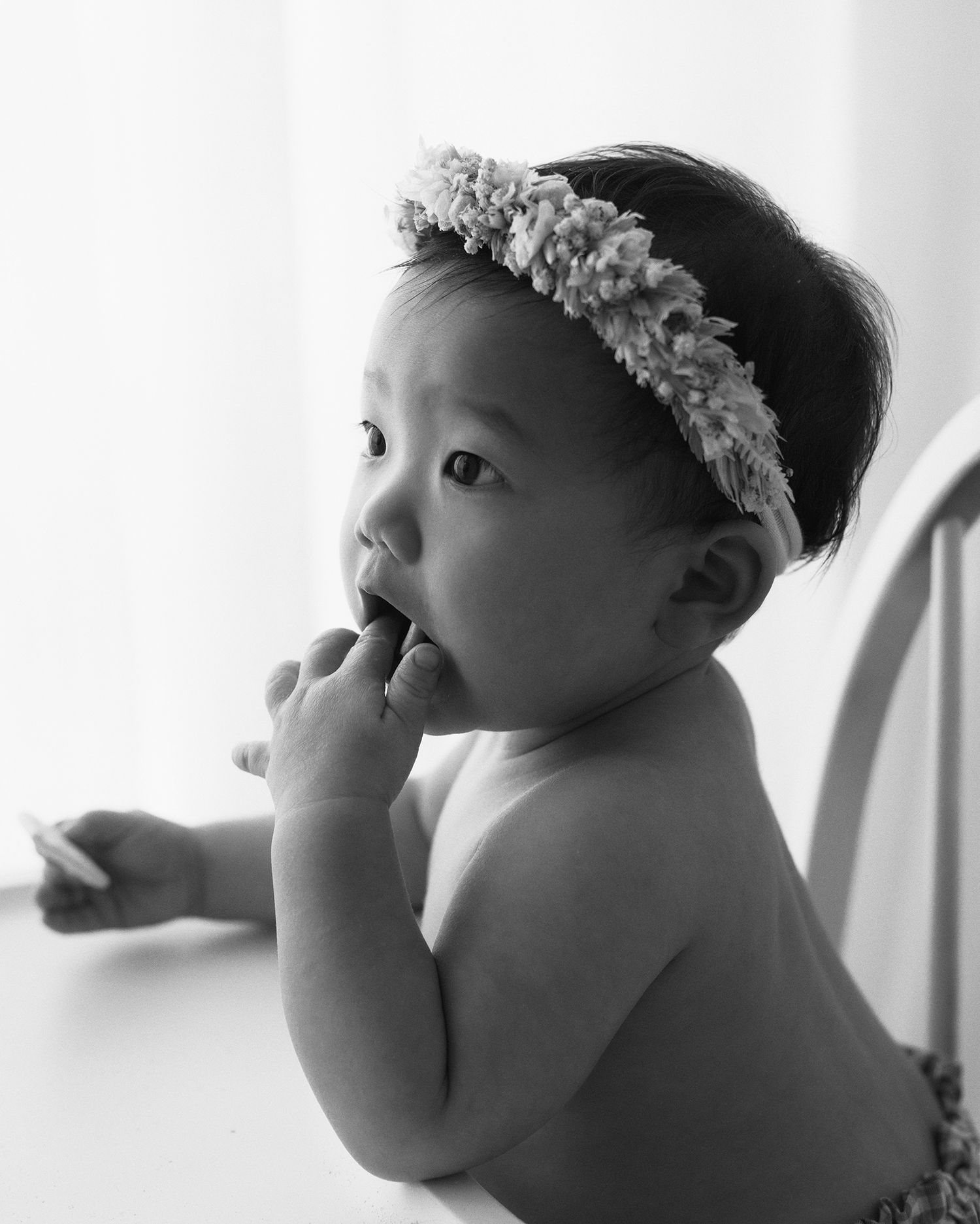 Black and white portrait of a baby in a flower crown chewing on their fingers while sitting at a high chair tray.