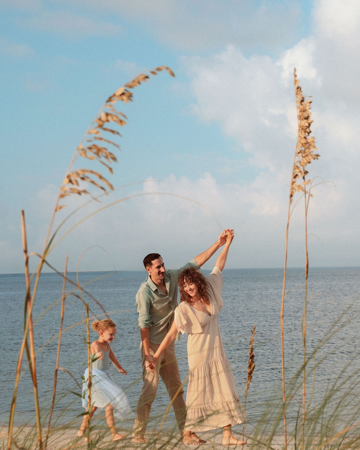 Family of three dancing and laughing on a sandy beach with tall sea oats, calm blue water, and a soft cloudy sky in the background.