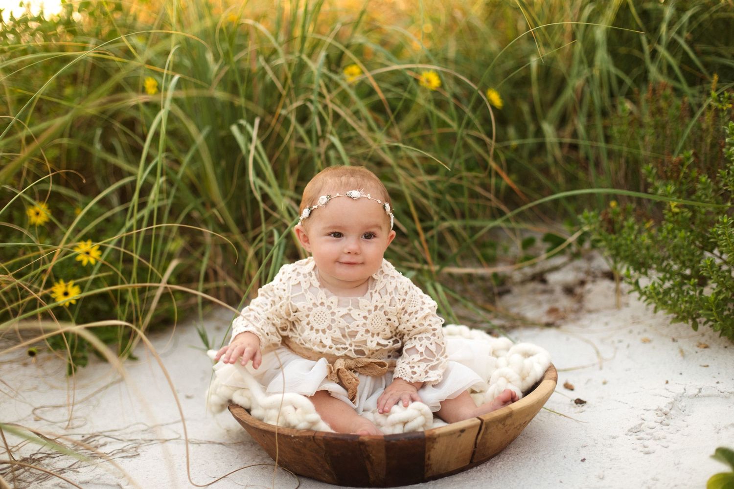 Smiling baby girl sitting in wooden bowl on white sand beach surrounded by green grass and yellow wildflowers.