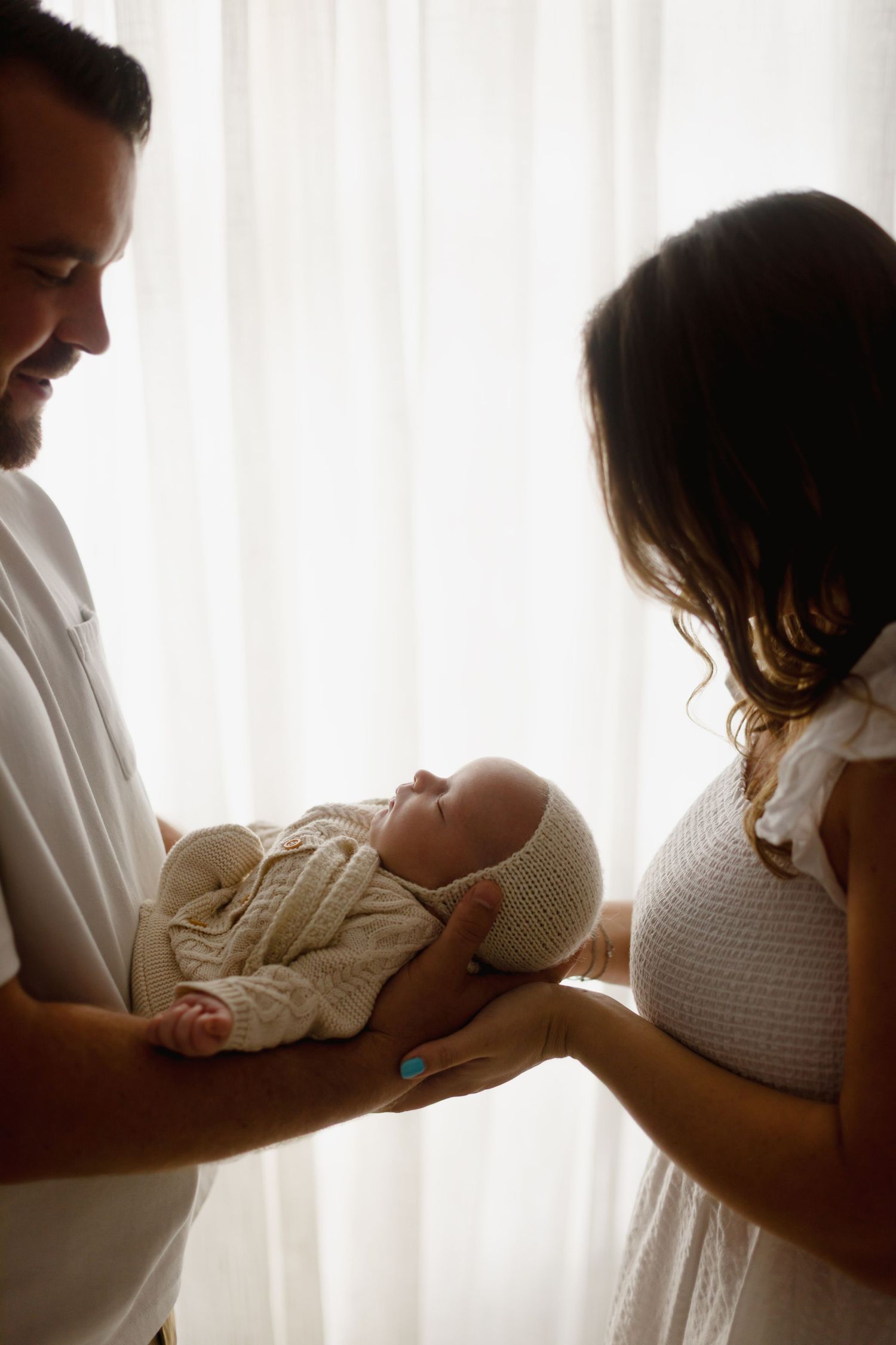 Smiling parents holding their sleeping newborn baby wrapped in cream knit outfit near a bright window.