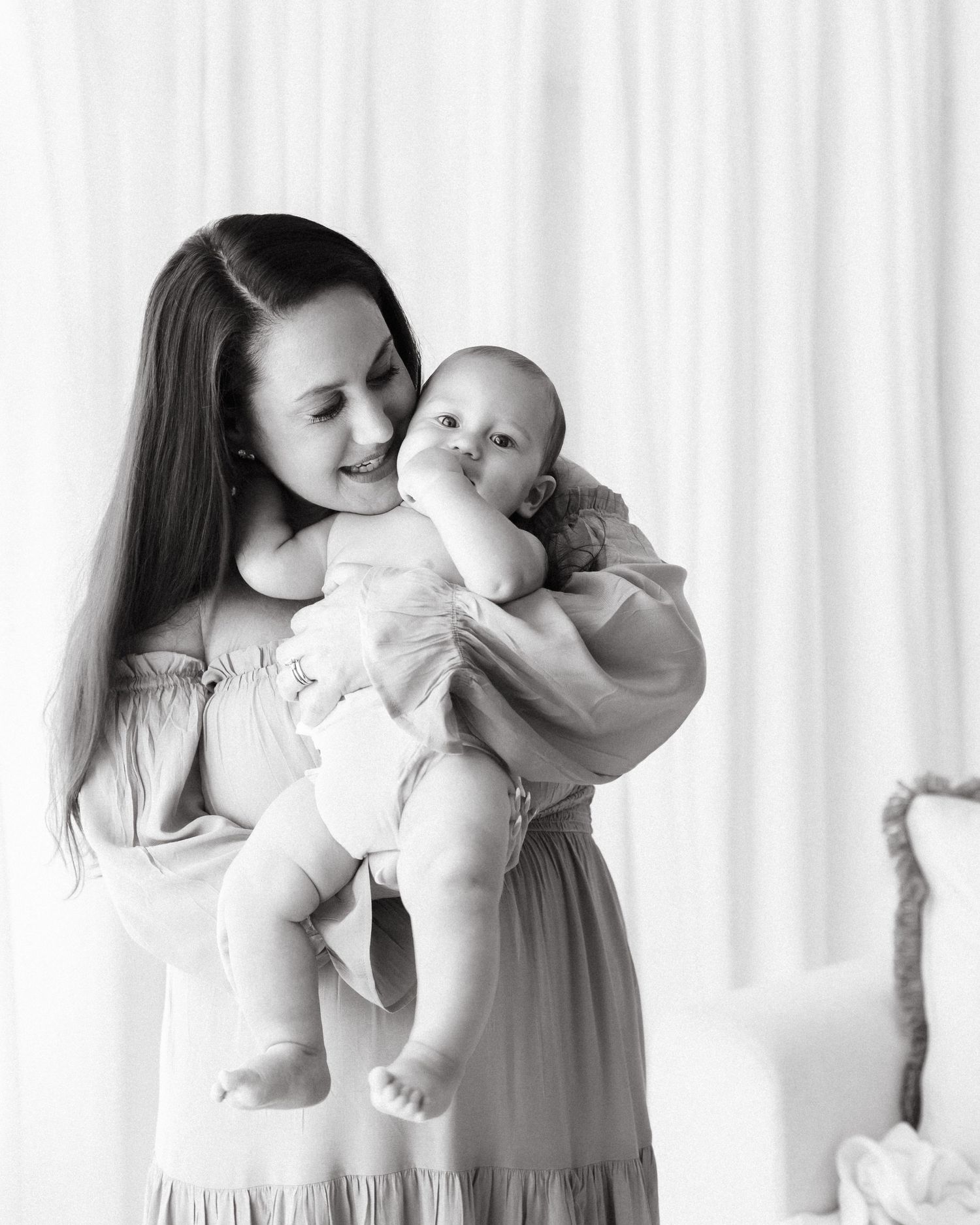 Black and white photo of a smiling mother in a flowy dress lovingly holding her happy baby in a bright studio setting.
