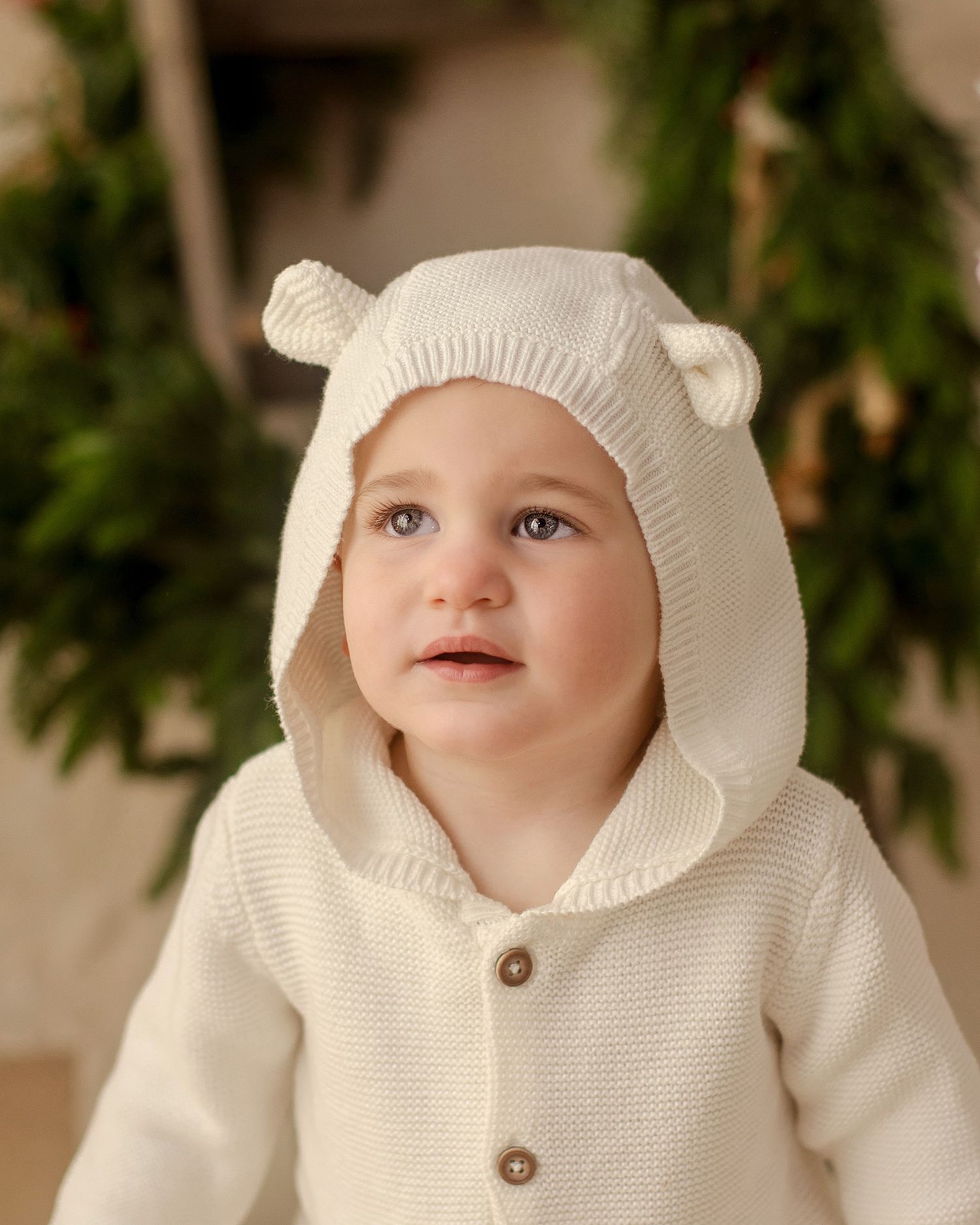 Adorable baby in cream knit bear-ear hooded sweater with wooden buttons, posed in front of festive green garland.