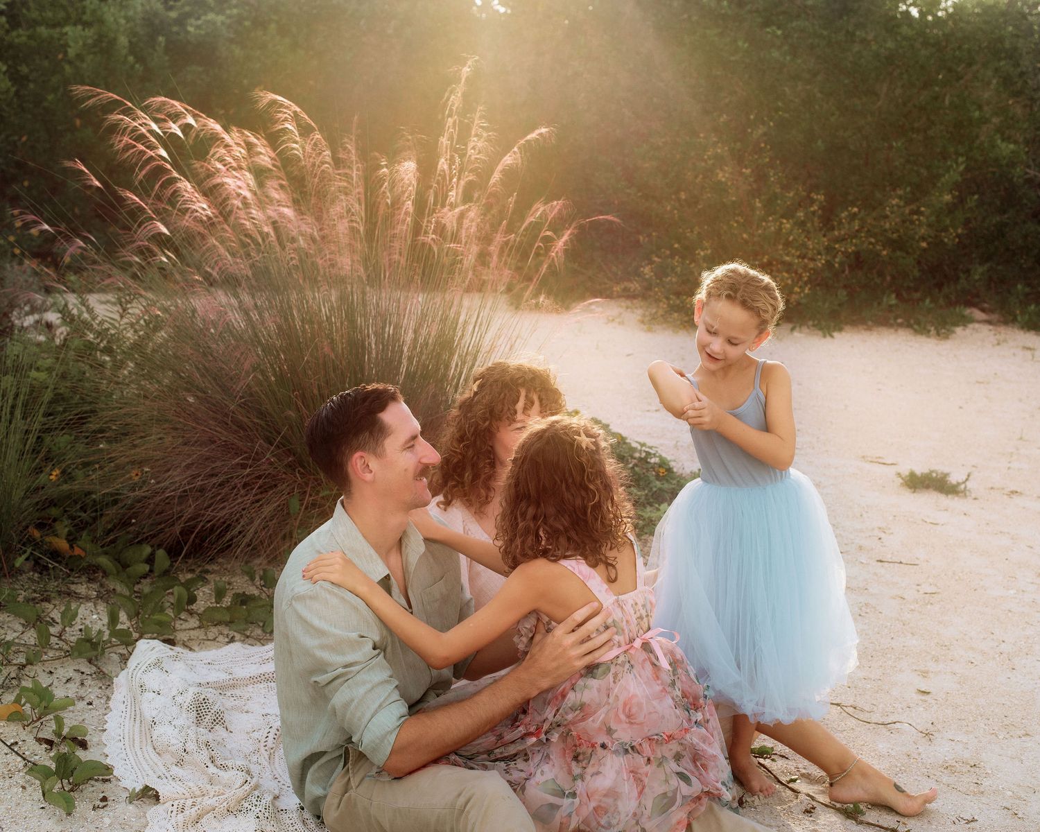 Family of four embracing on a sandy beach at golden hour, with a girl in a blue tutu skirt standing nearby beside tall pink grasses.