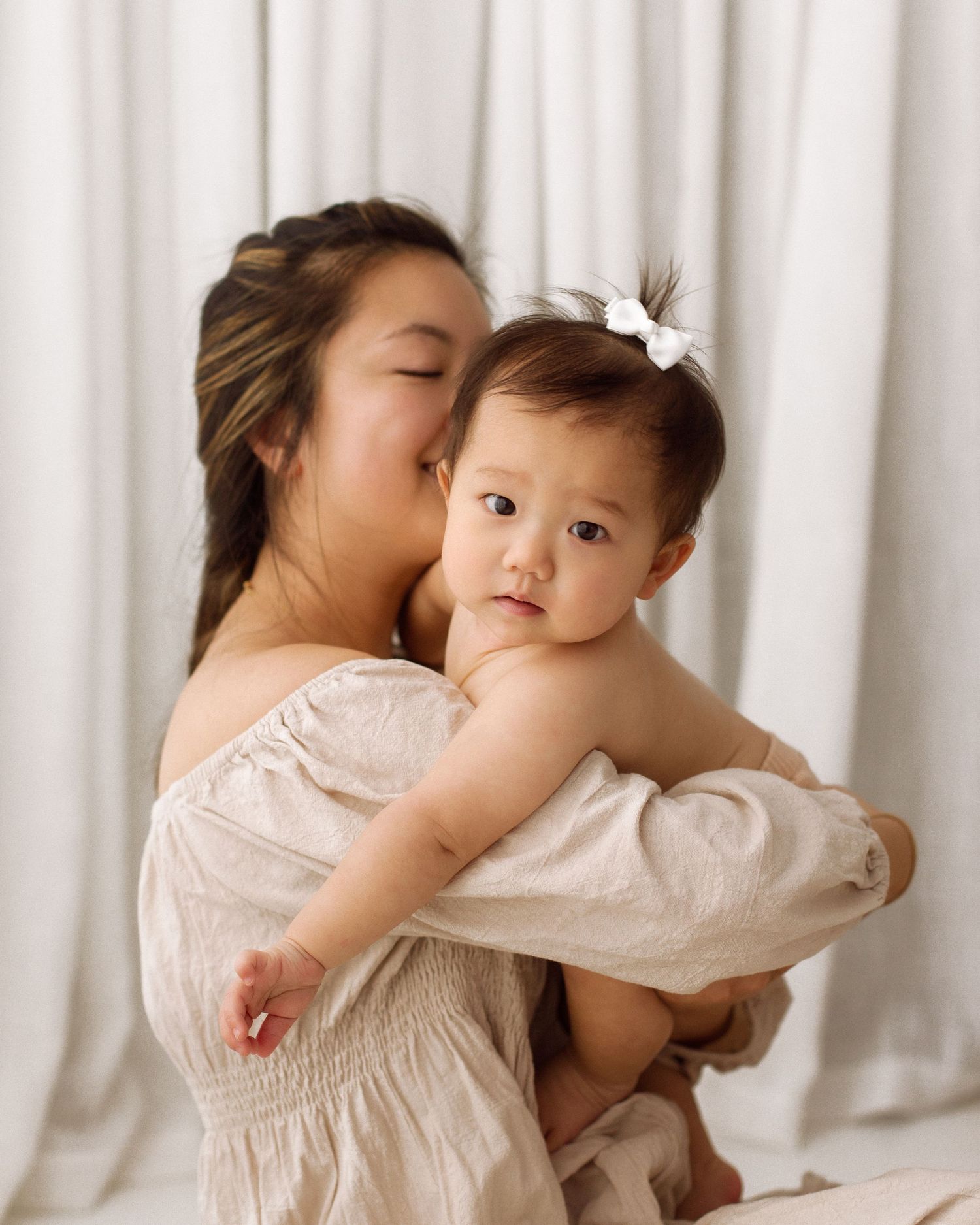 A woman in a cream off-the-shoulder dress holds a baby with a white bow in her hair against a soft white curtain backdrop.