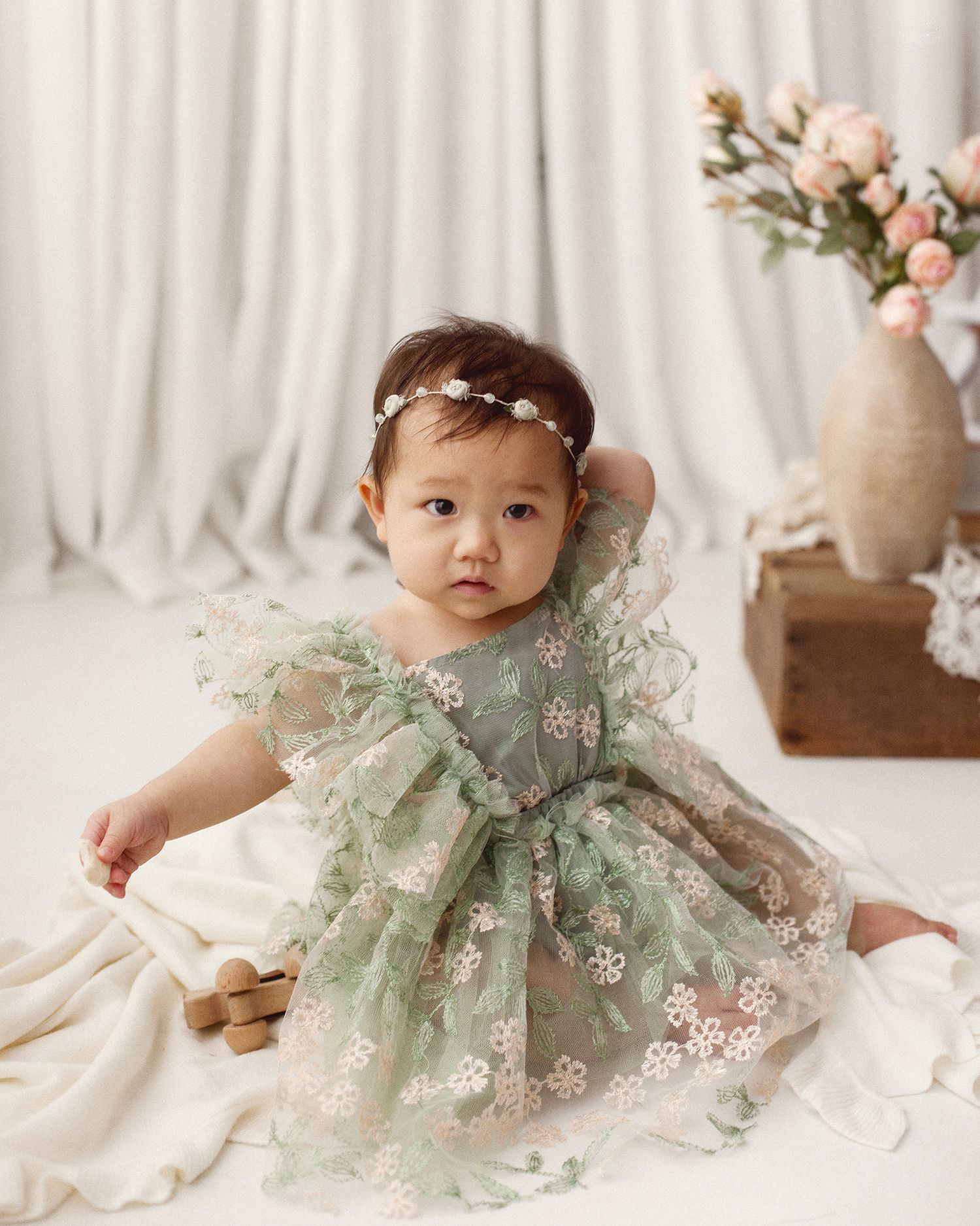 Baby girl in a sage green floral embroidered tulle dress and flower headband sitting on a cream blanket beside pink roses in a vase.