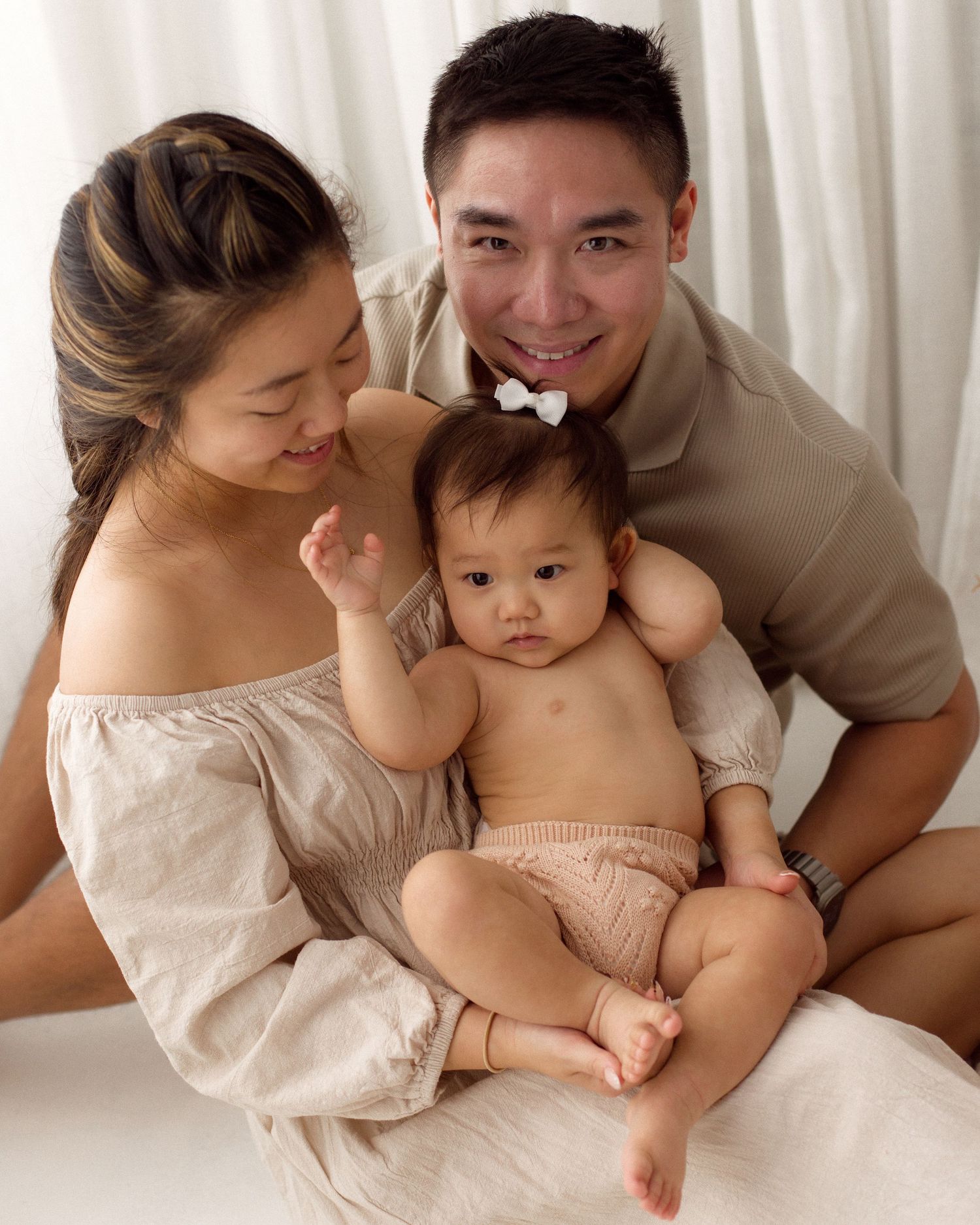 Smiling mother and father in neutral tones hold their baby girl with a white hair bow in a bright, soft studio family portrait.