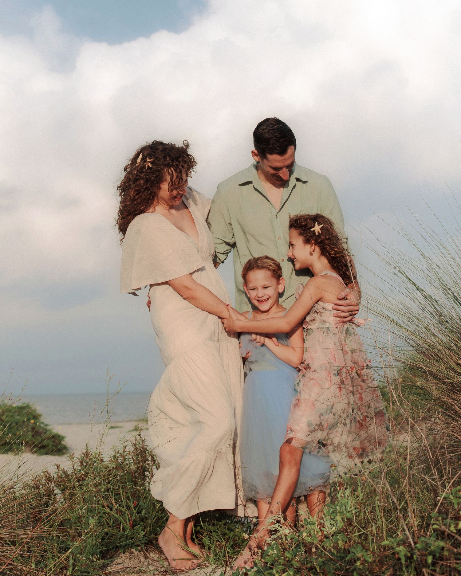 Family of four embracing in beach dunes with sea oats, wearing flowing neutral outfits under a soft cloudy sky at the shore.