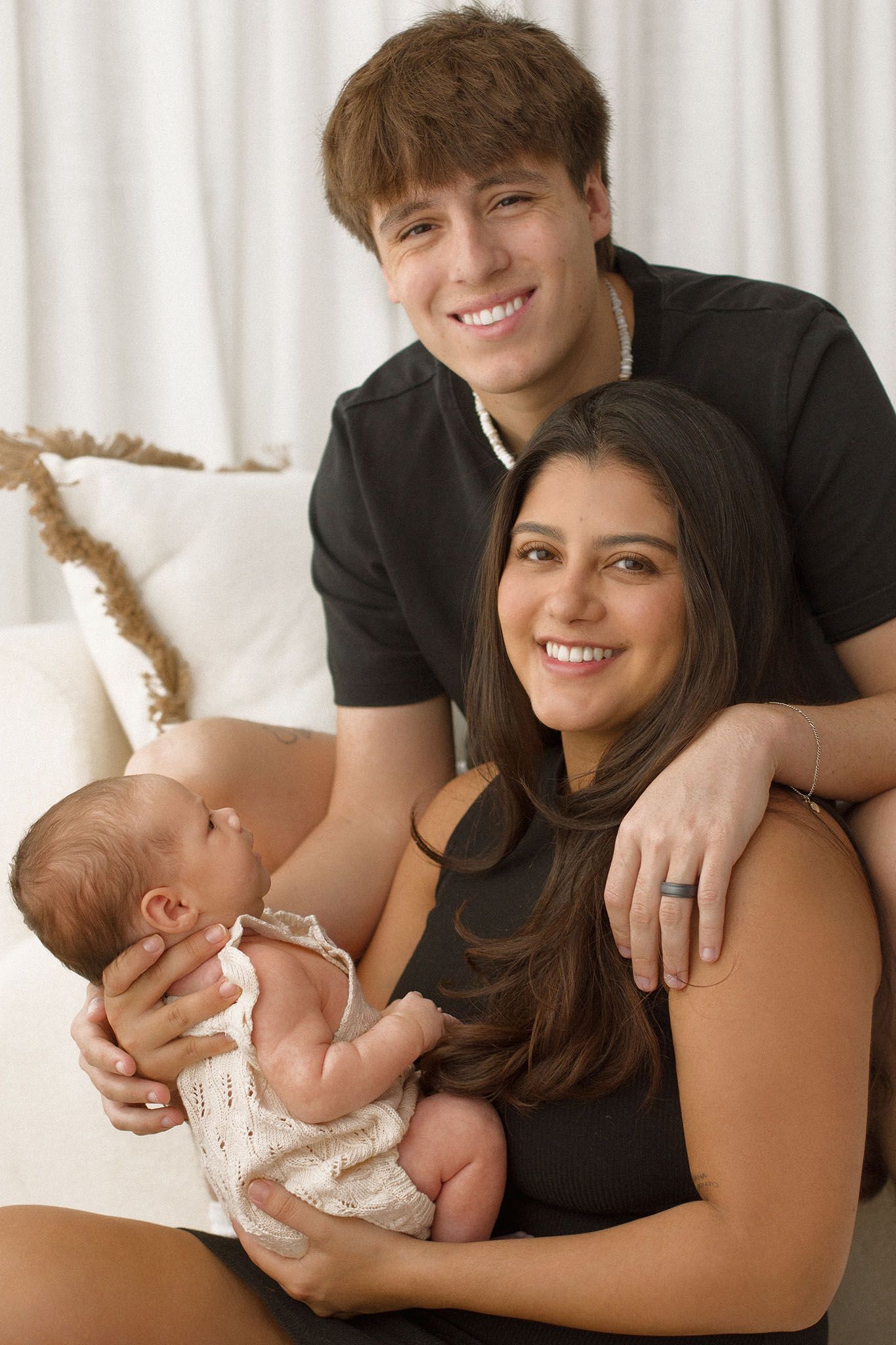 Young couple in black outfits smiling while holding their newborn baby at home.
