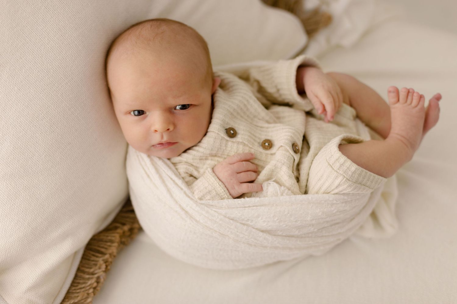 Wide-eyed newborn baby swaddled in cream knit wrap with button cardigan on white backdrop.