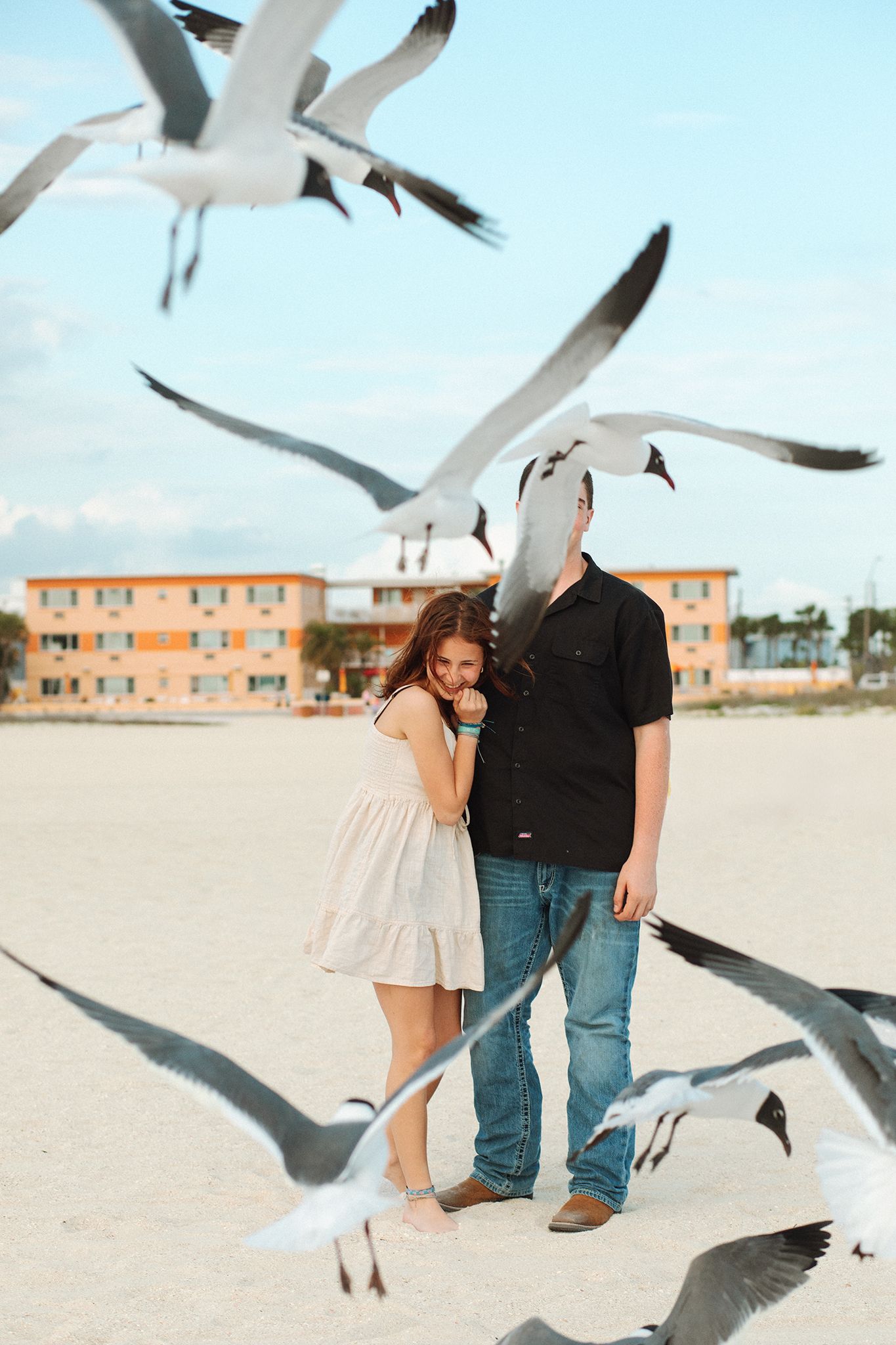 Couple embracing on sandy beach surrounded by flying seagulls, with a colorful building in the background.