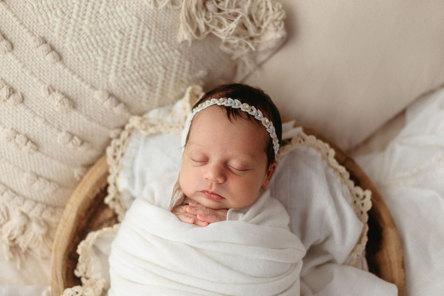 Newborn baby girl swaddled in white wrap with floral headband, sleeping peacefully in a wooden bowl prop.