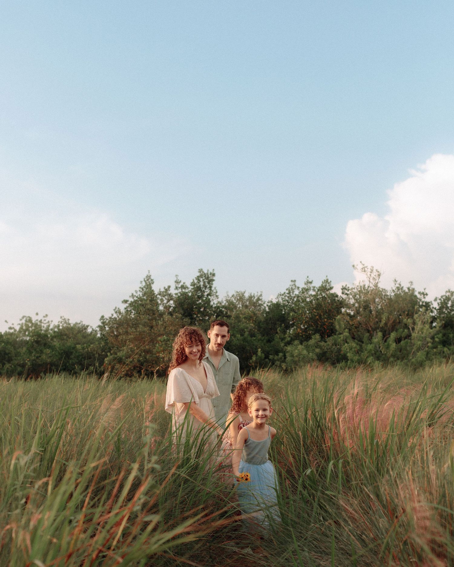 Family of four standing together in tall golden grass under a blue sky with soft clouds, shot during a warm outdoor portrait session.