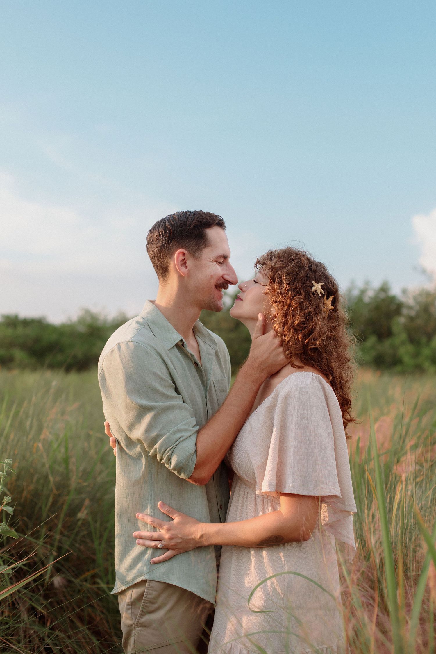 Smiling couple embracing and touching noses in a tall golden grass field at sunset, in an outdoor engagement or couples photoshoot.
