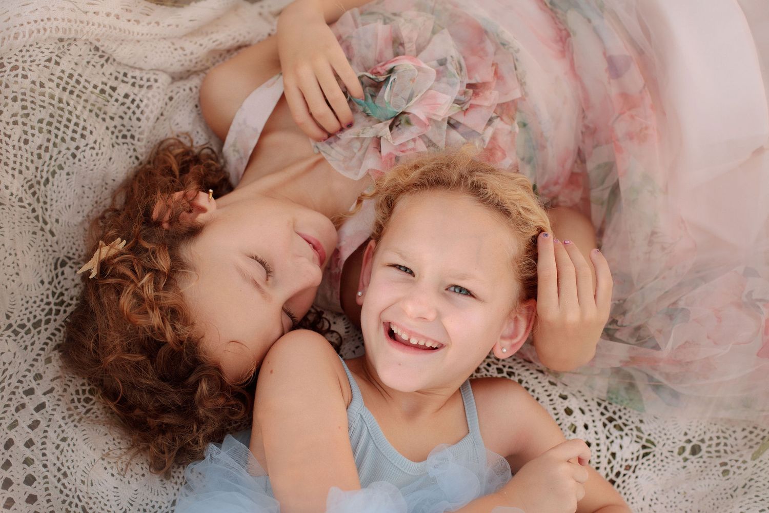Two smiling young sisters in soft pastel tulle dresses cuddle together on a cream lace blanket in a sweet overhead portrait.