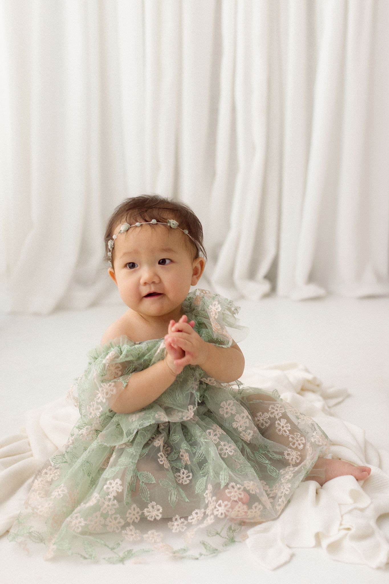 Baby girl in a green floral tulle dress and beaded headband sits with hands clasped on a cream blanket in front of white curtains.