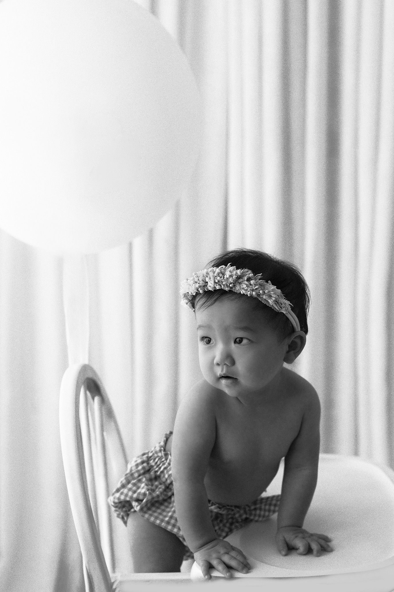 Black and white photo of a baby in a floral headband and checkered bottoms climbing on a white chair beside a large balloon.
