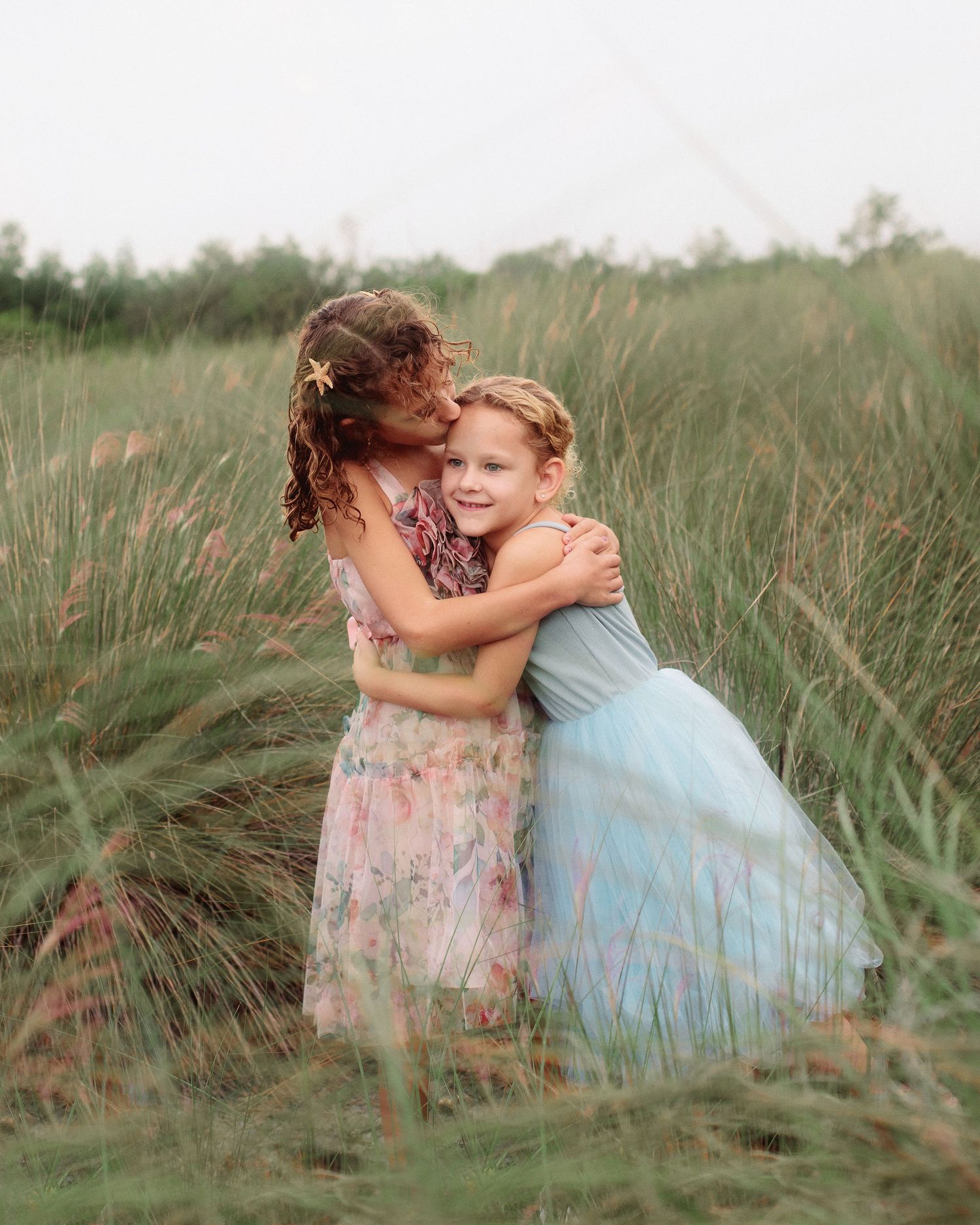 Two young girls in floral and light blue tulle dresses share a loving hug in a tall grass meadow.