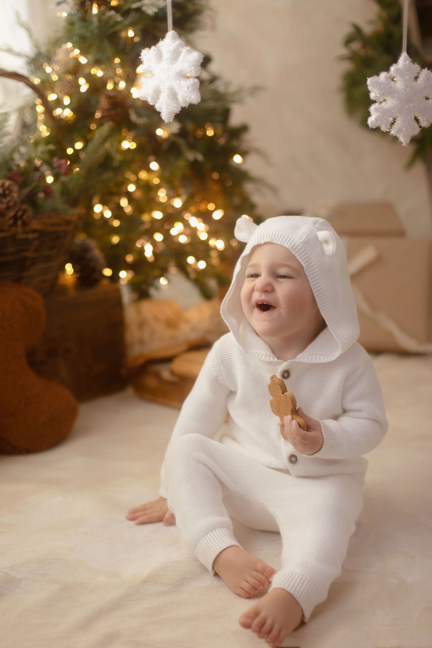 Happy baby in white bear onesie sits on fur rug by lit Christmas tree holding a gingerbread cookie.