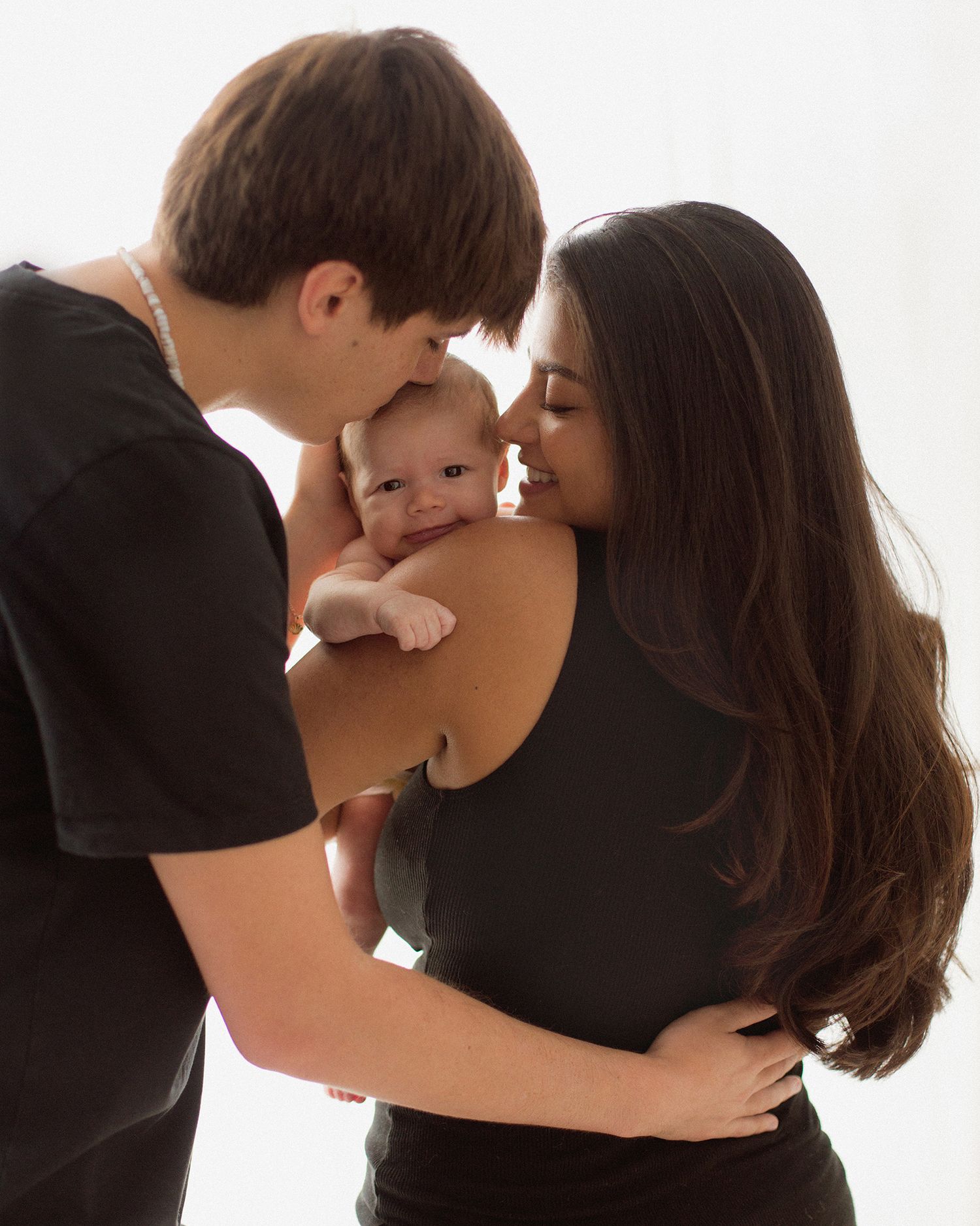 Young couple in black outfits lovingly holding and kissing their smiling newborn baby against white background.