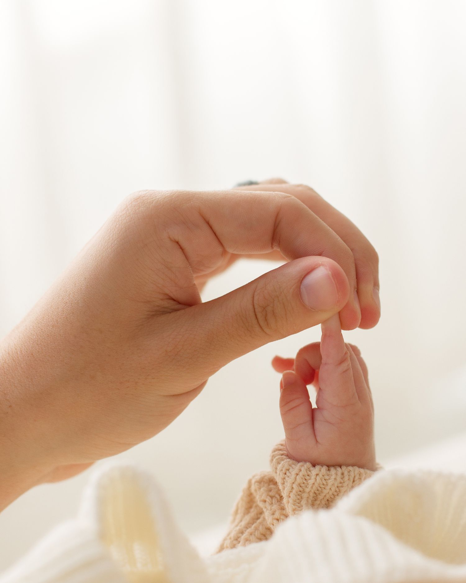 A gentle adult hand holding a tiny newborn baby's finger, wrapped in a soft knit sleeve, against a white background.
