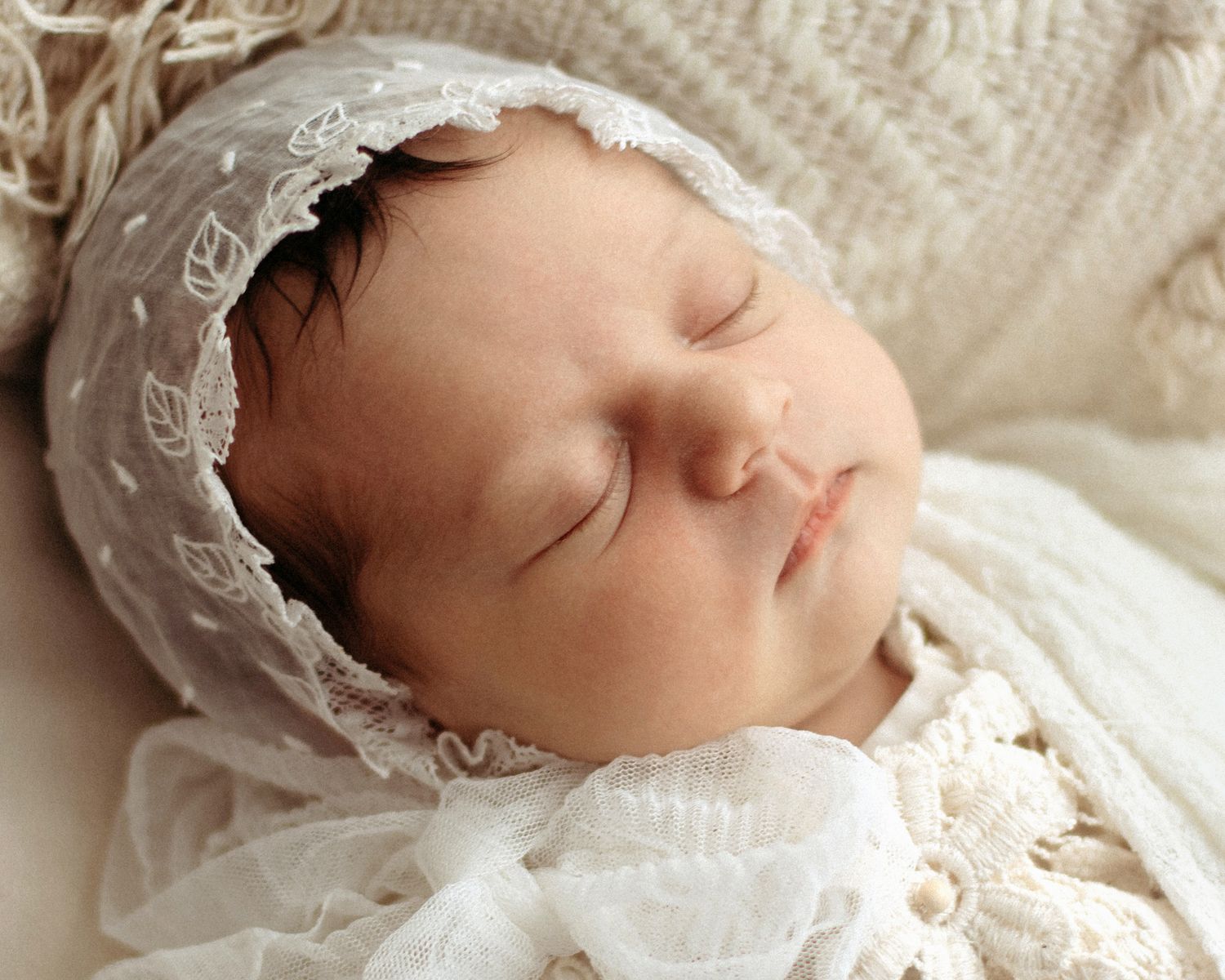 Peaceful newborn baby sleeping in white lace bonnet and swaddle wrap, soft portrait photography.