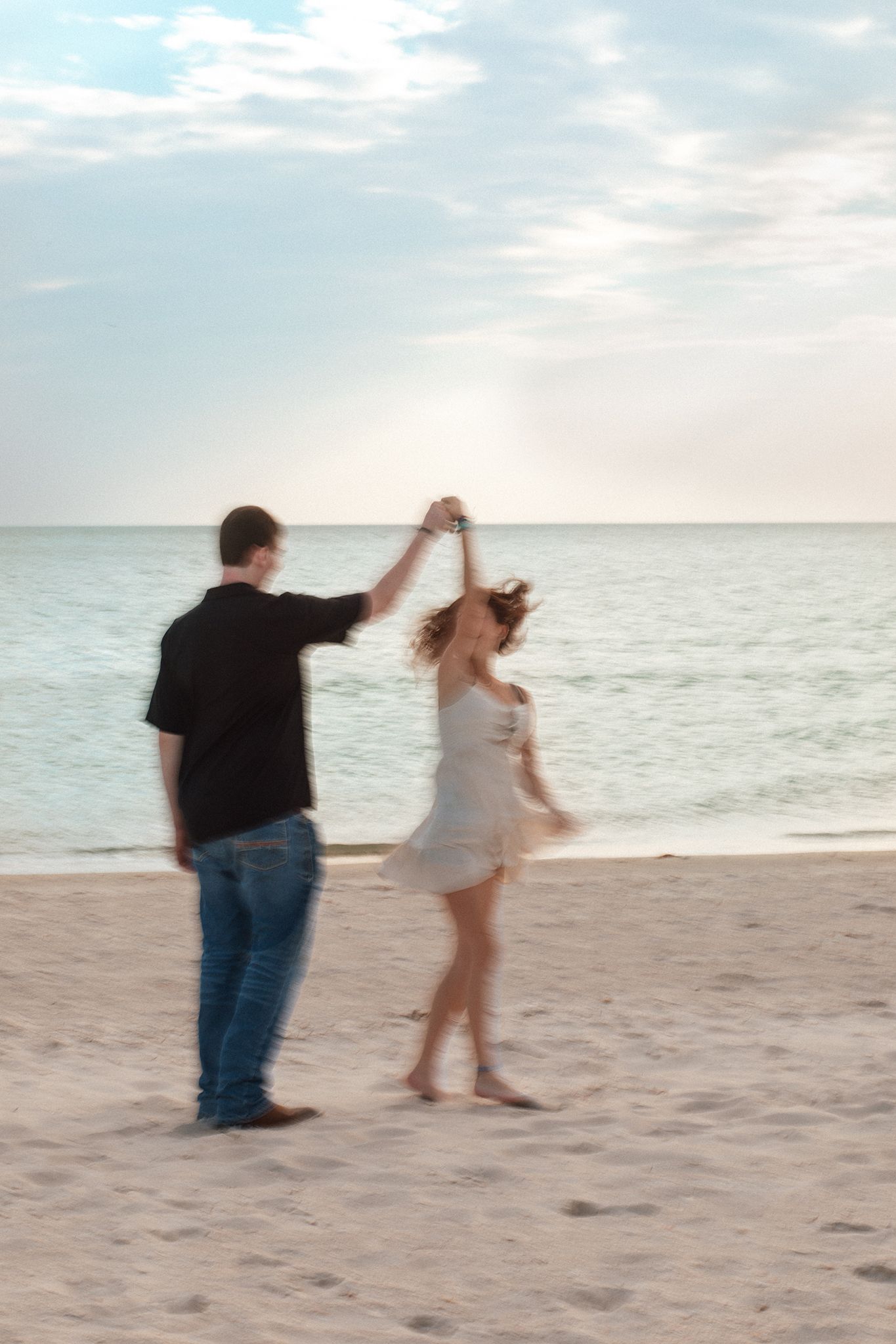 Couple dancing romantically on a sandy beach at sunset, man in black shirt twirling woman in white dress.