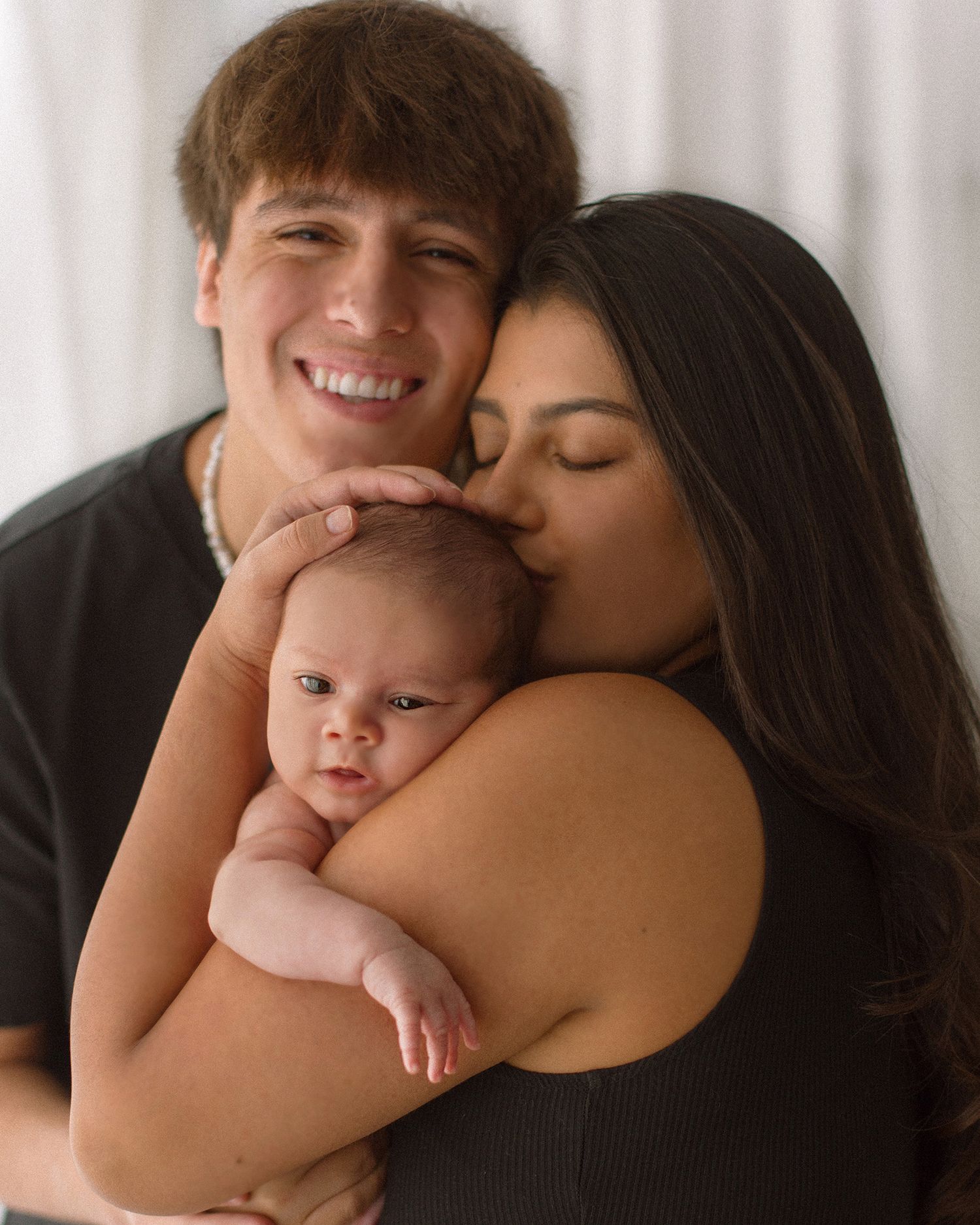A young couple smiling and kissing their newborn baby during a professional family portrait session.