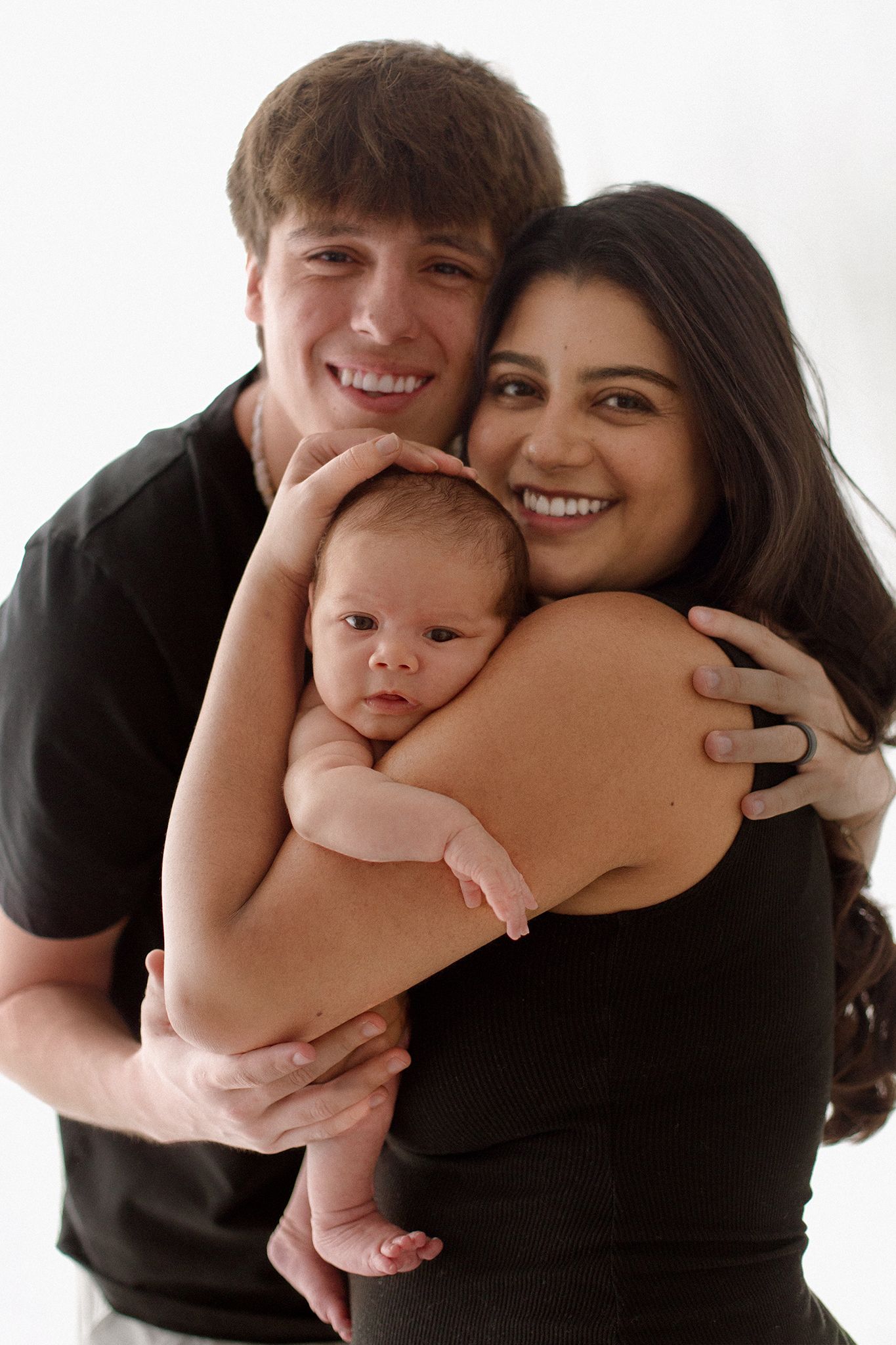 Smiling young couple in black shirts holding their newborn baby against a white background.