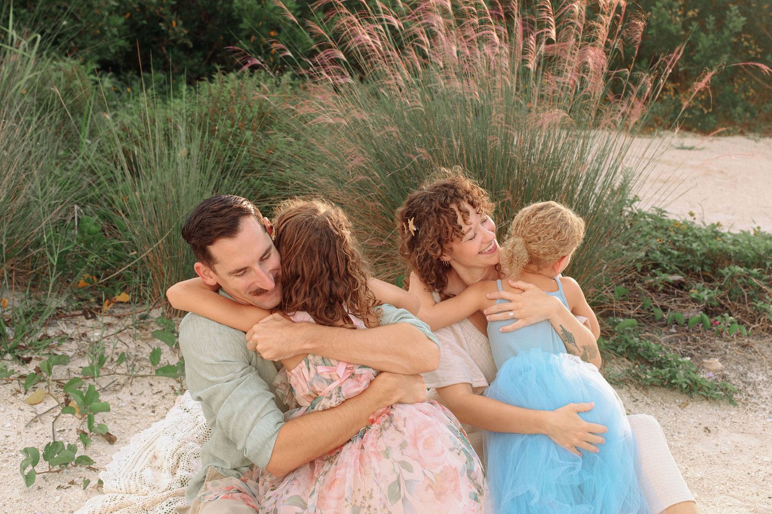 A family of four hugs closely outdoors among tall coastal grasses, with two girls in pastel dresses embraced by their smiling parents.