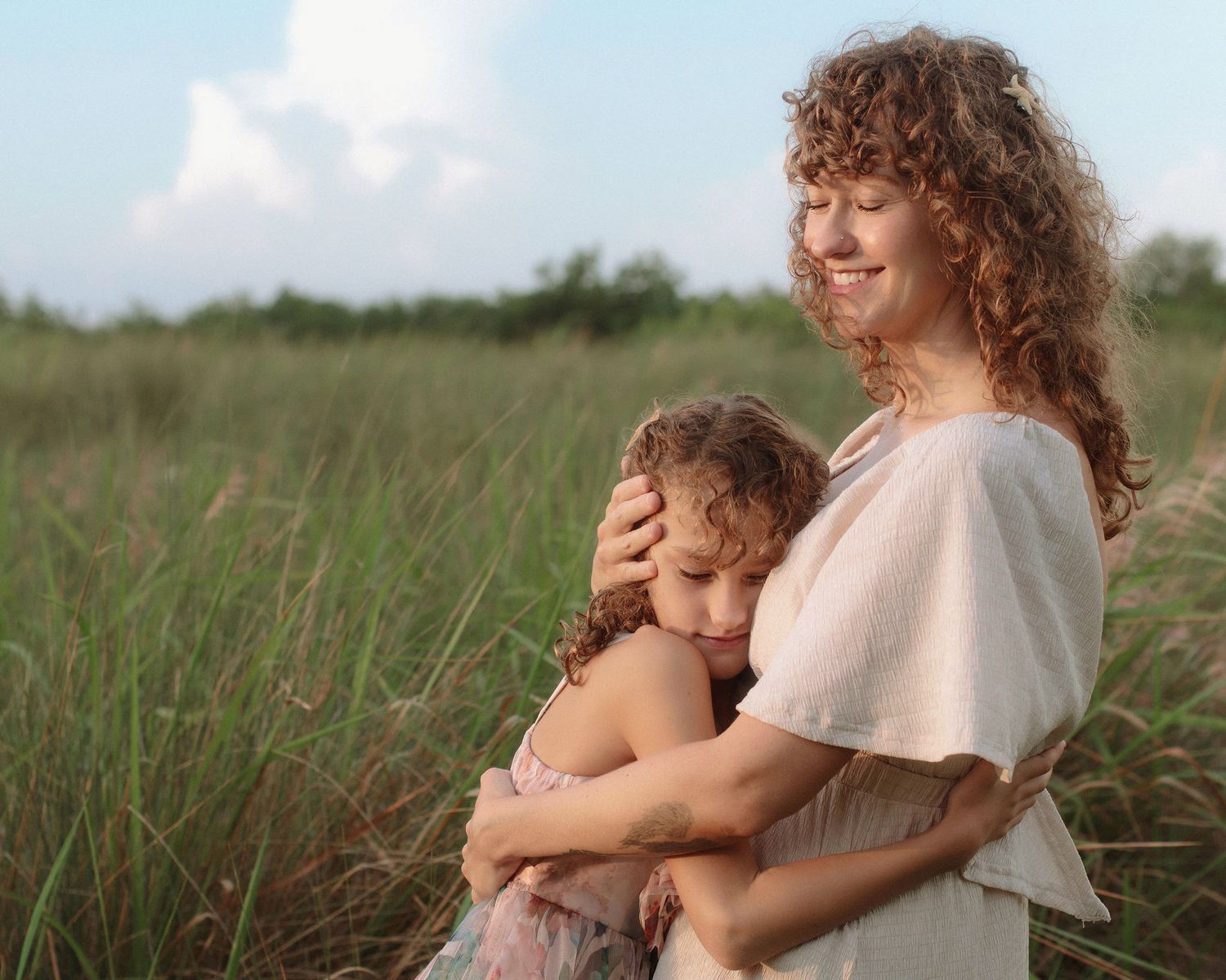 Smiling woman with curly hair hugs her young daughter in a tall green grass field under a bright blue sky.