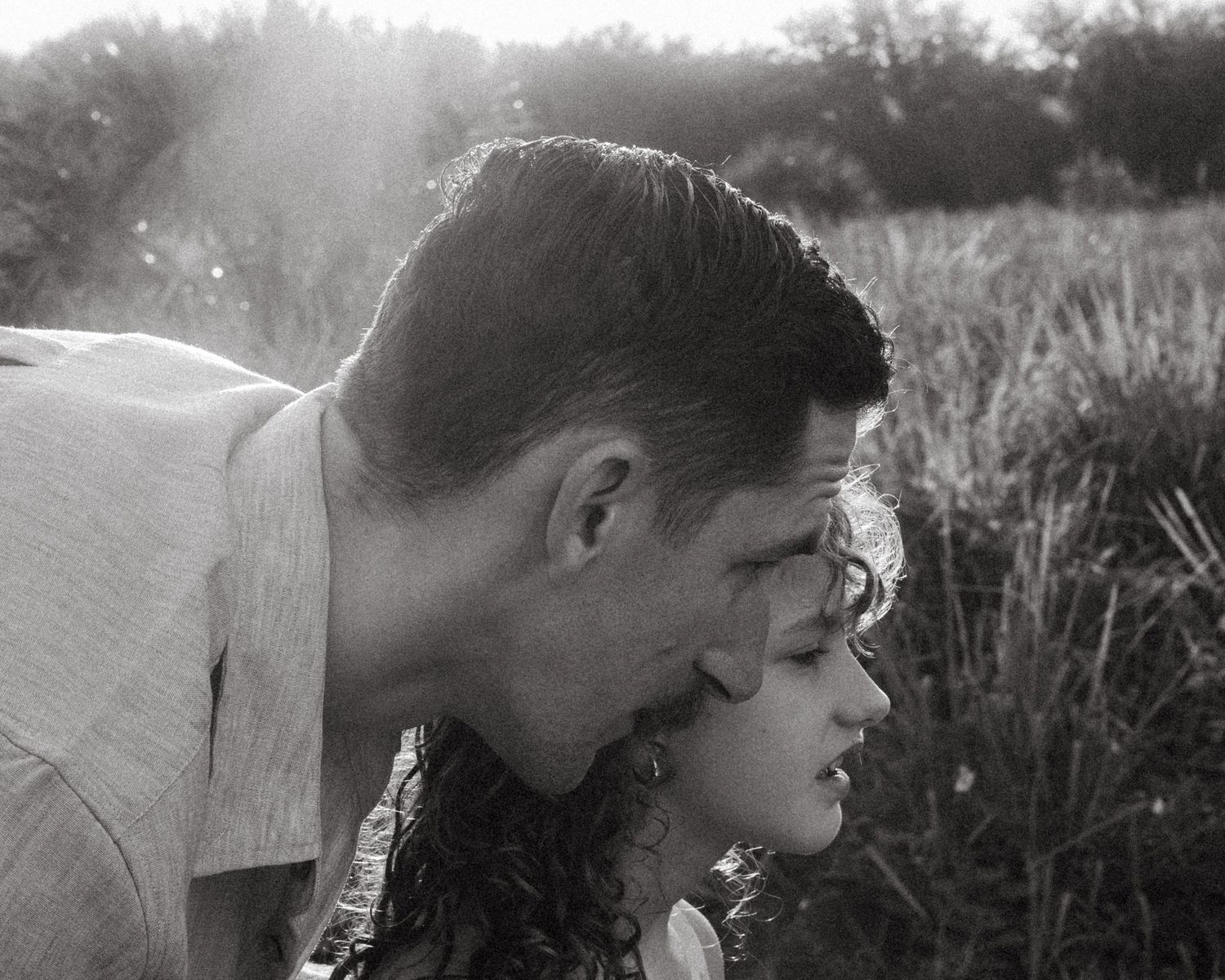 Black and white photo of a man leaning in to kiss a curly-haired woman beside tall grass in soft, hazy sunlight.