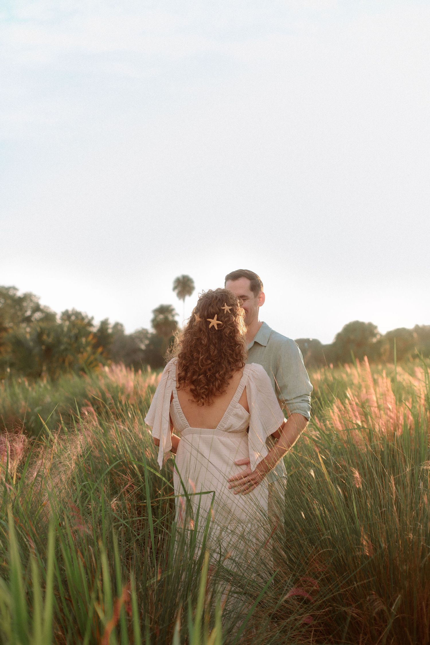 Couple embracing in tall golden grass at sunset, the woman in a white dress with starfish hair accessories and curly hair.