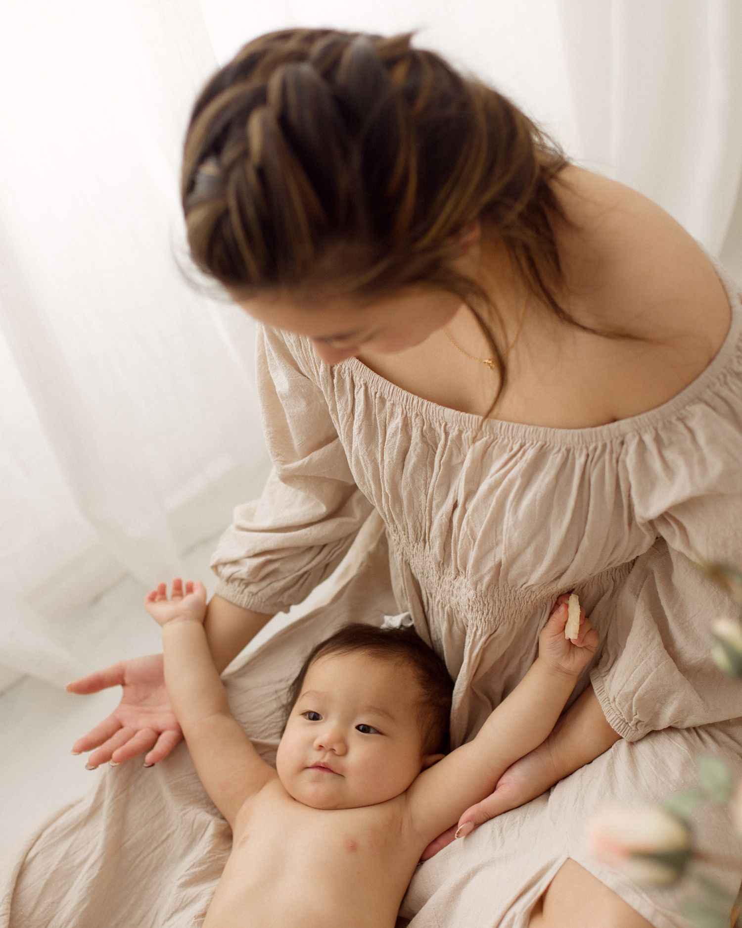 Mother in a beige off-shoulder dress with a braided hairstyle sits and gently holds her smiling baby in a bright, soft-lit studio.